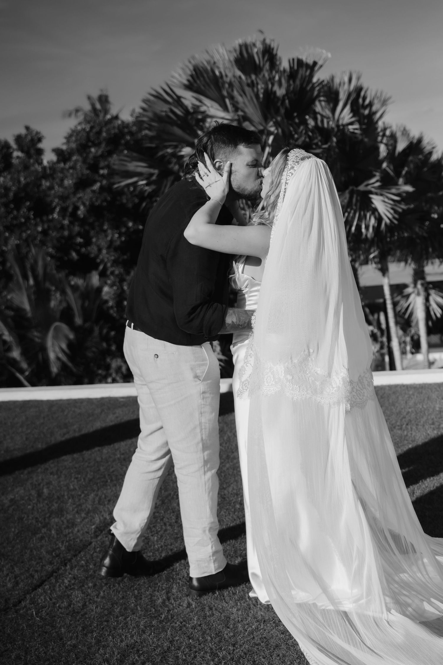 Black and white photo of bride and groom sharing first kiss with tropical palm trees in the background.
