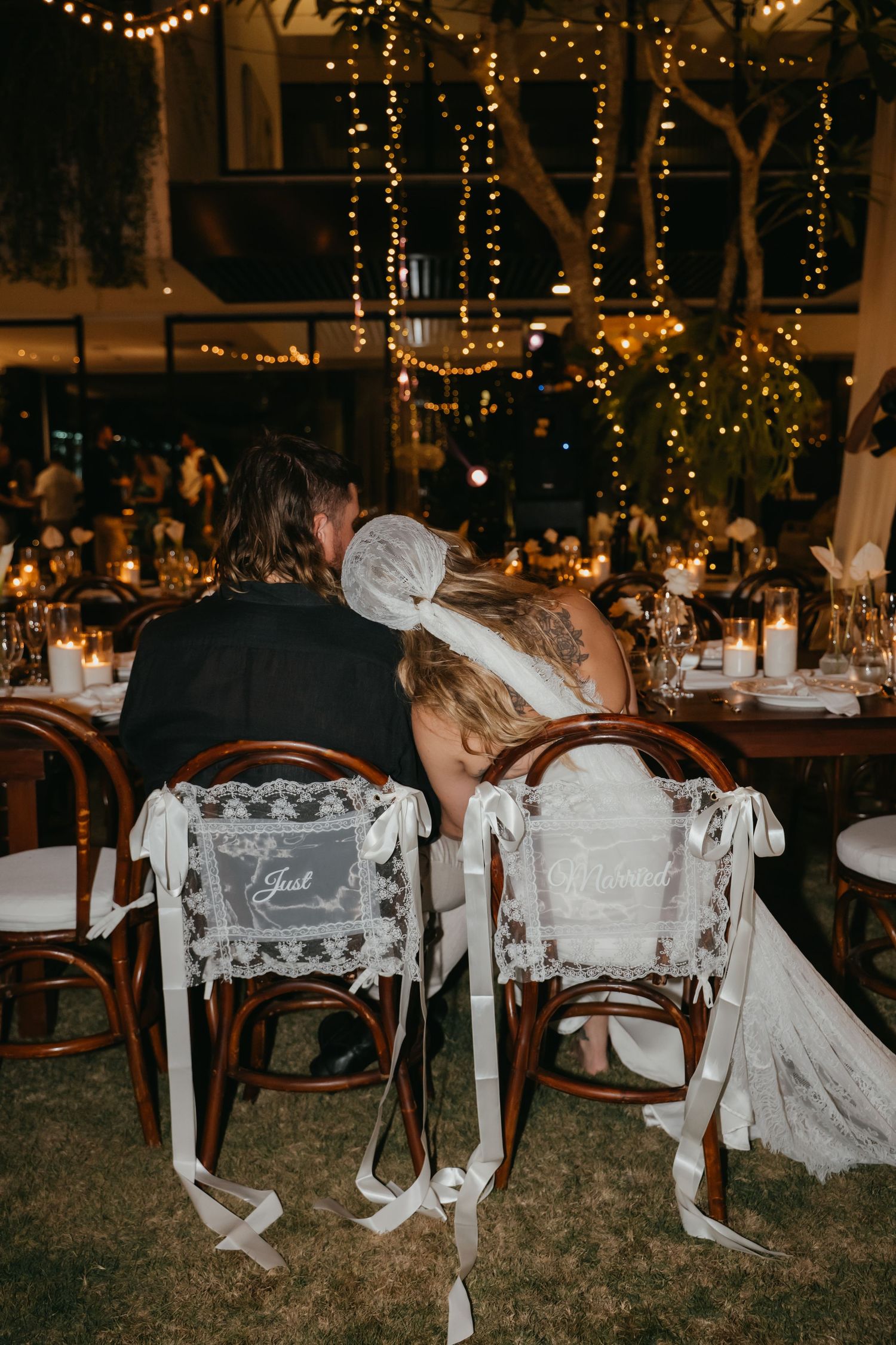 Bride and groom lean together at decorated reception table under warm string lights at night.