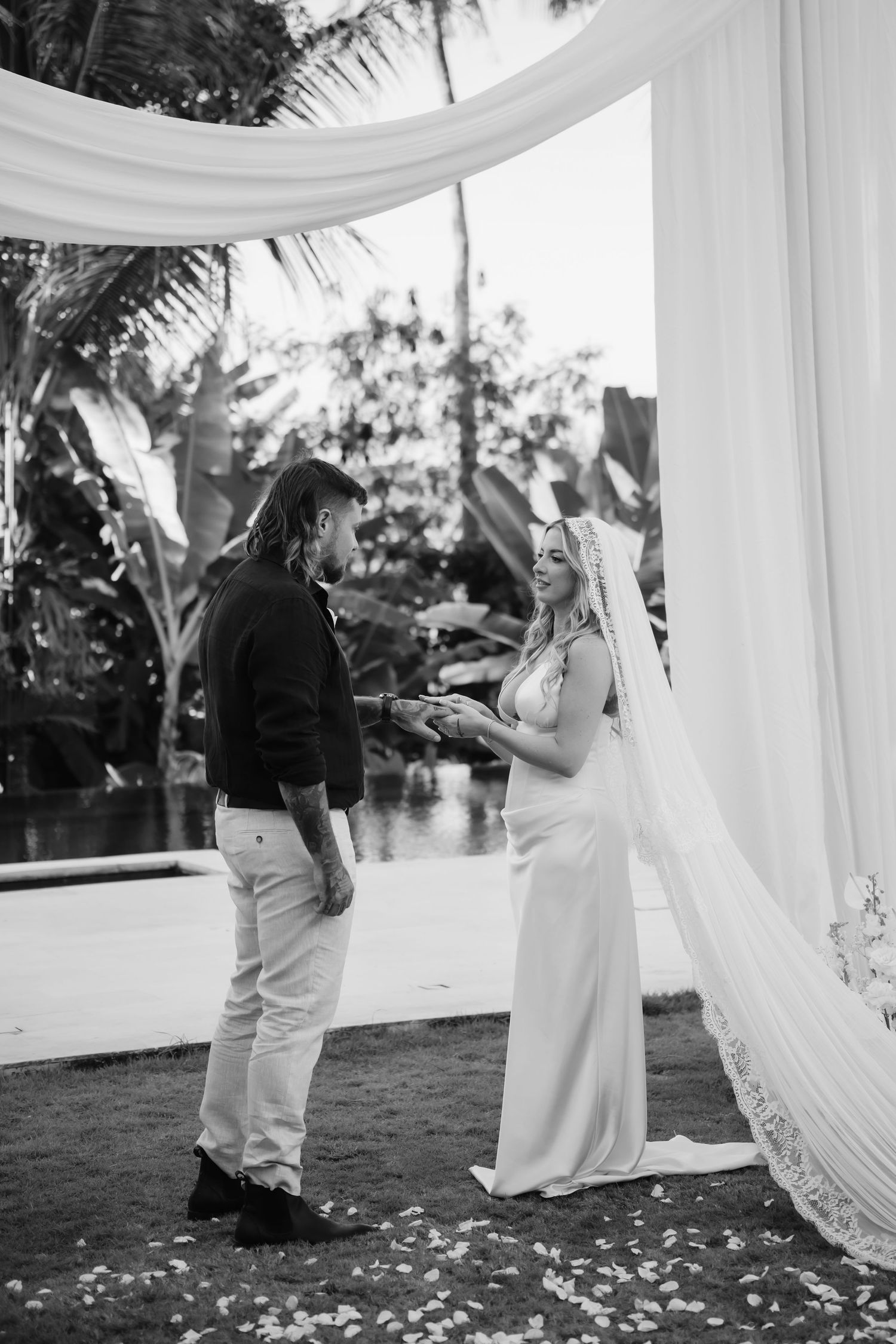 Bride and groom exchange vows under white draped wedding arch surrounded by tropical greenery in black and white photo.