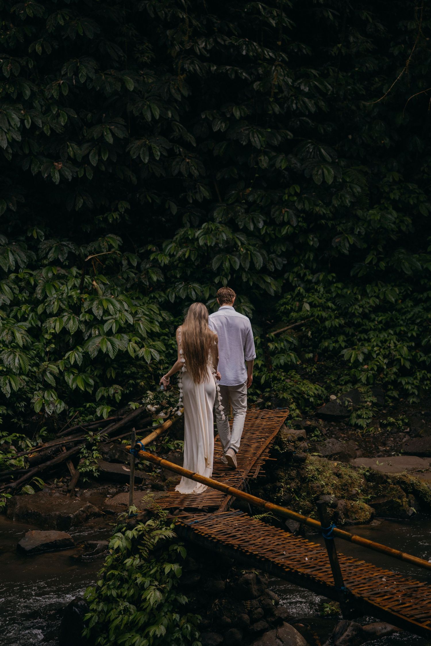 A couple in light clothing stands together on a wooden bridge over a river surrounded by lush green foliage.
