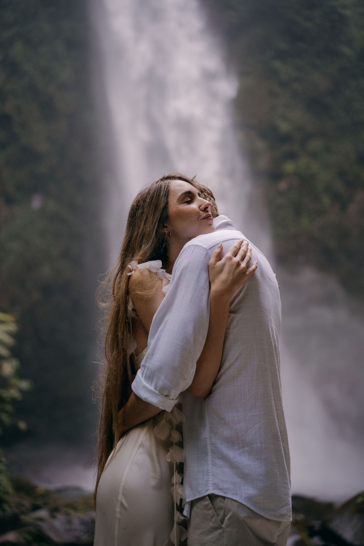A couple embraces in front of a misty waterfall in a romantic outdoor setting.