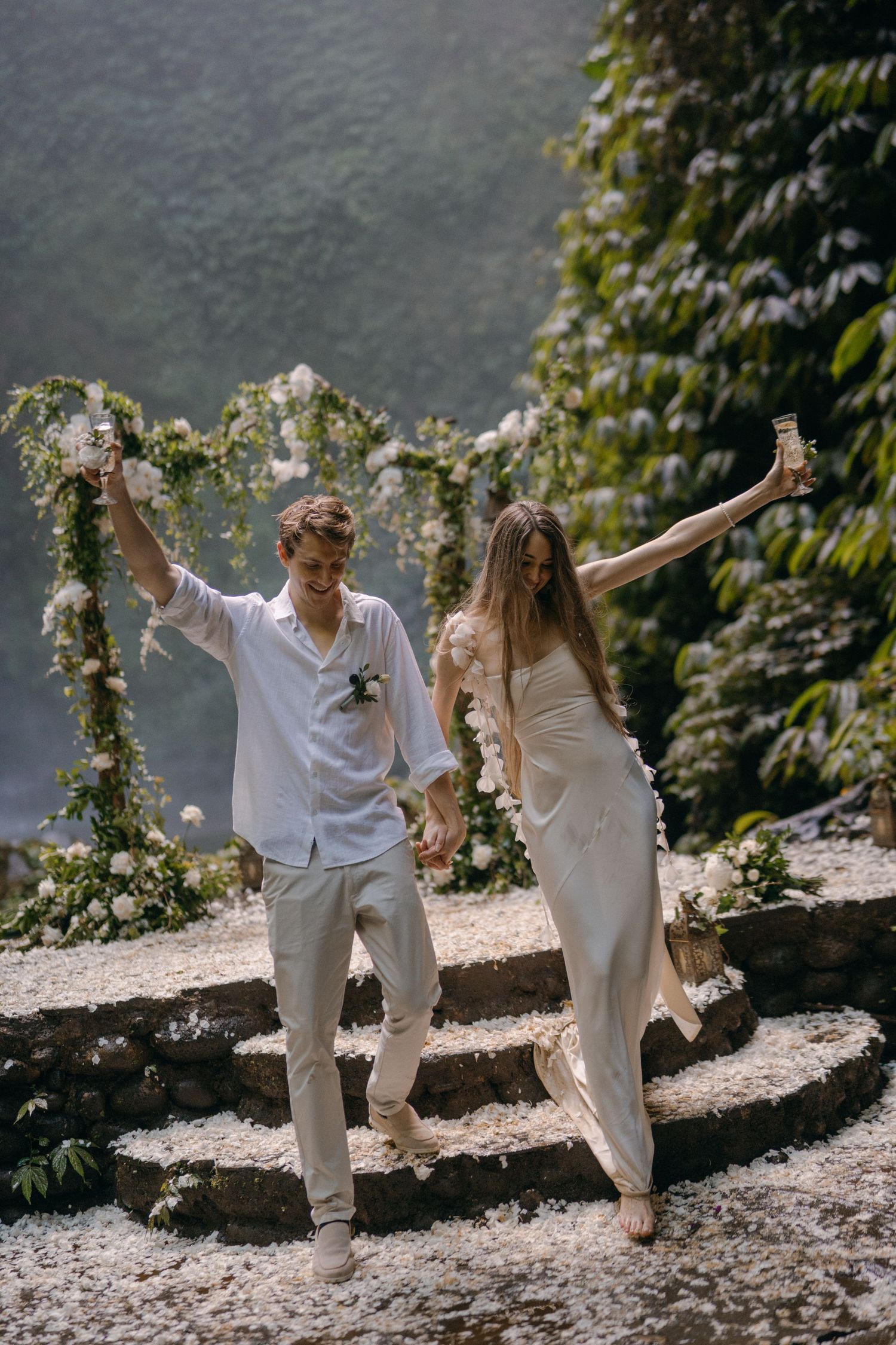 A couple in white outfits dances joyfully together on a sunlit path surrounded by greenery and flowers.