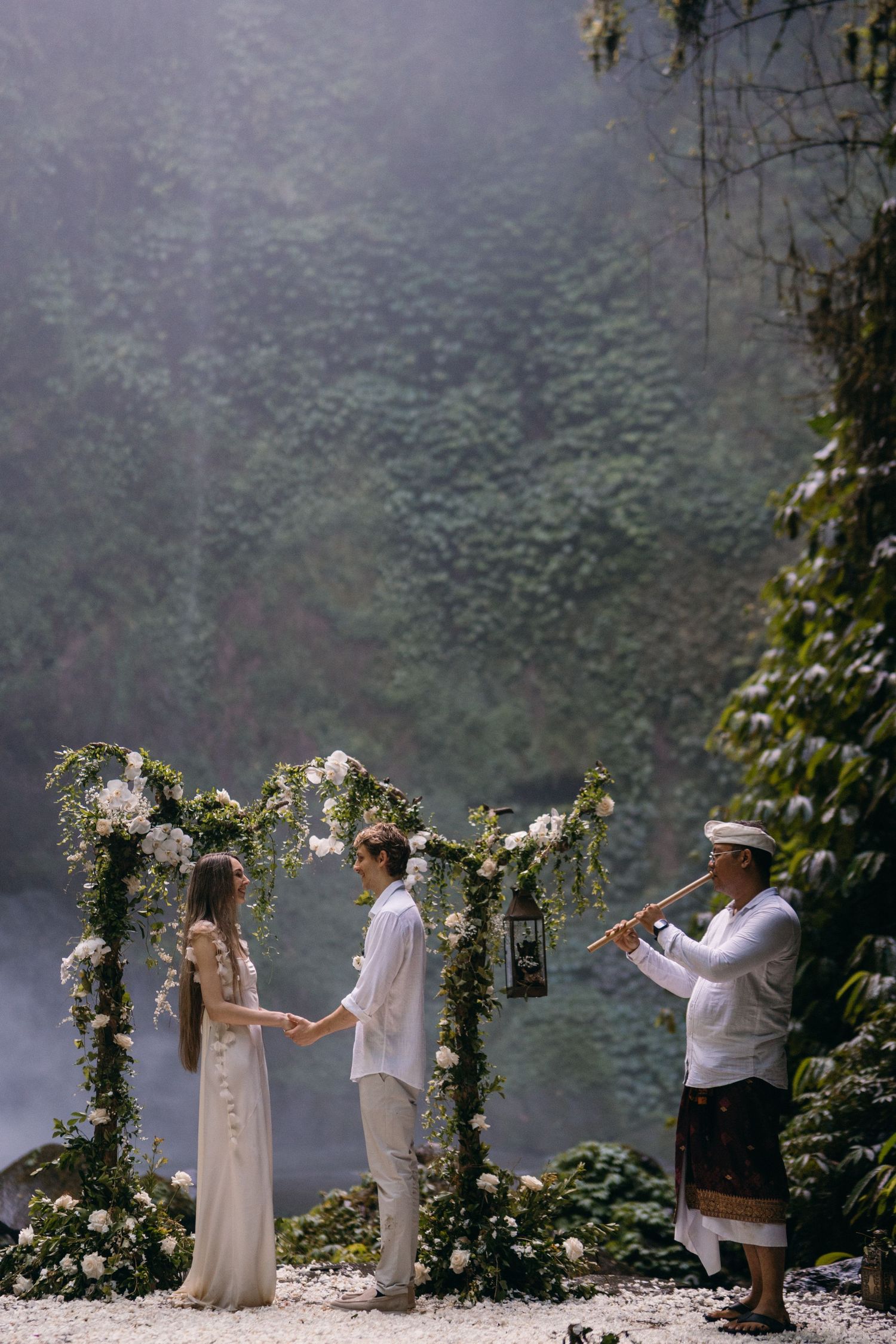 A romantic outdoor wedding ceremony under a floral arch with a violinist playing music in a misty forest setting.