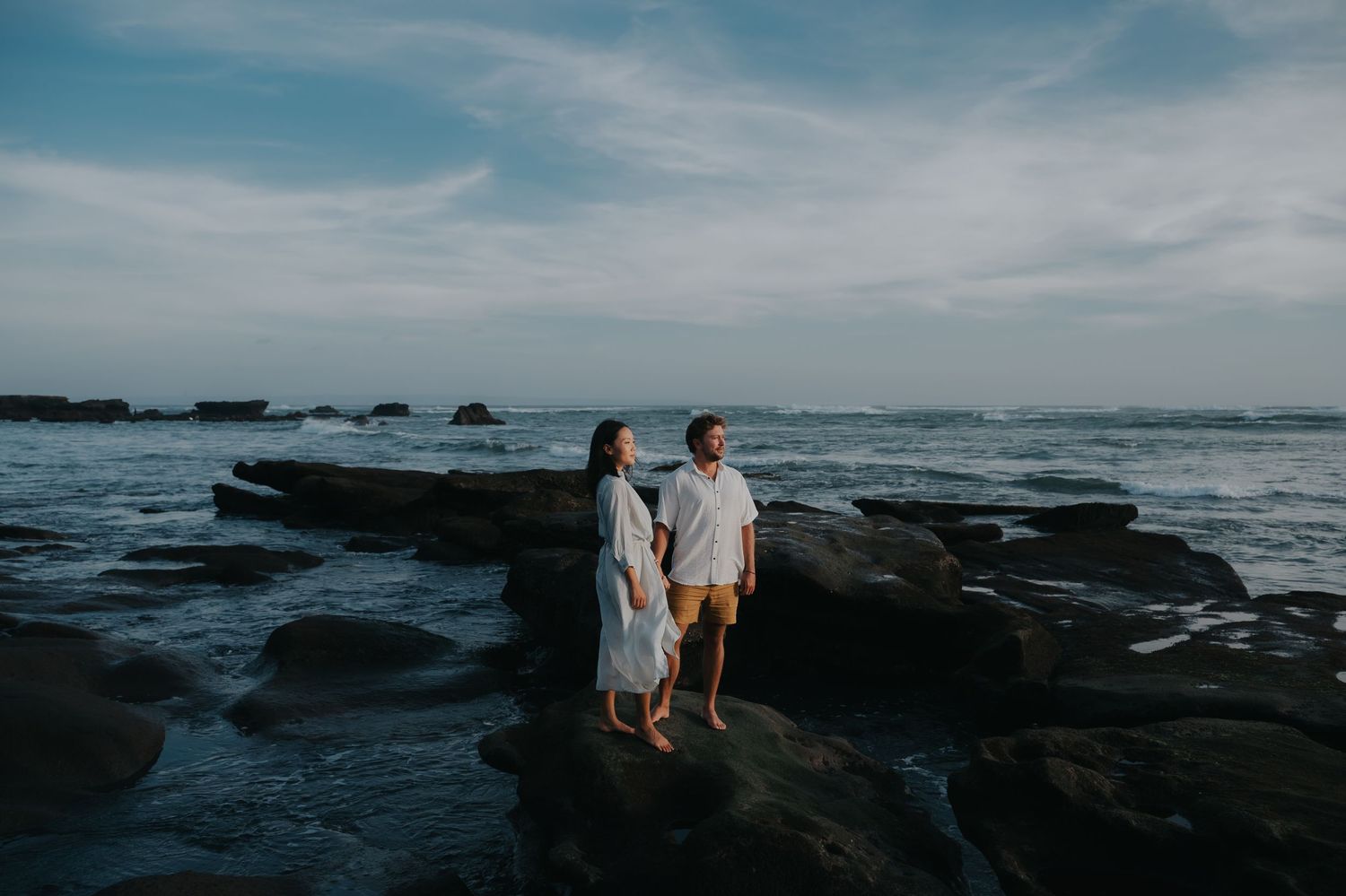 A couple stands on rocky coastal cliffs overlooking the ocean at sunset creating a romantic silhouette against the sky.