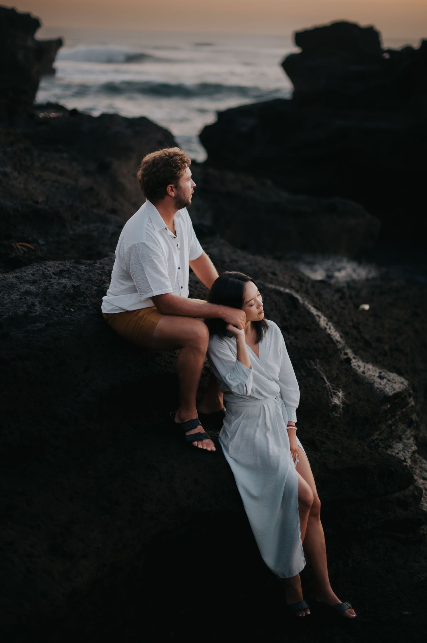 A couple sits together on dark rocky cliffs at sunset overlooking the ocean in romantic silhouette.