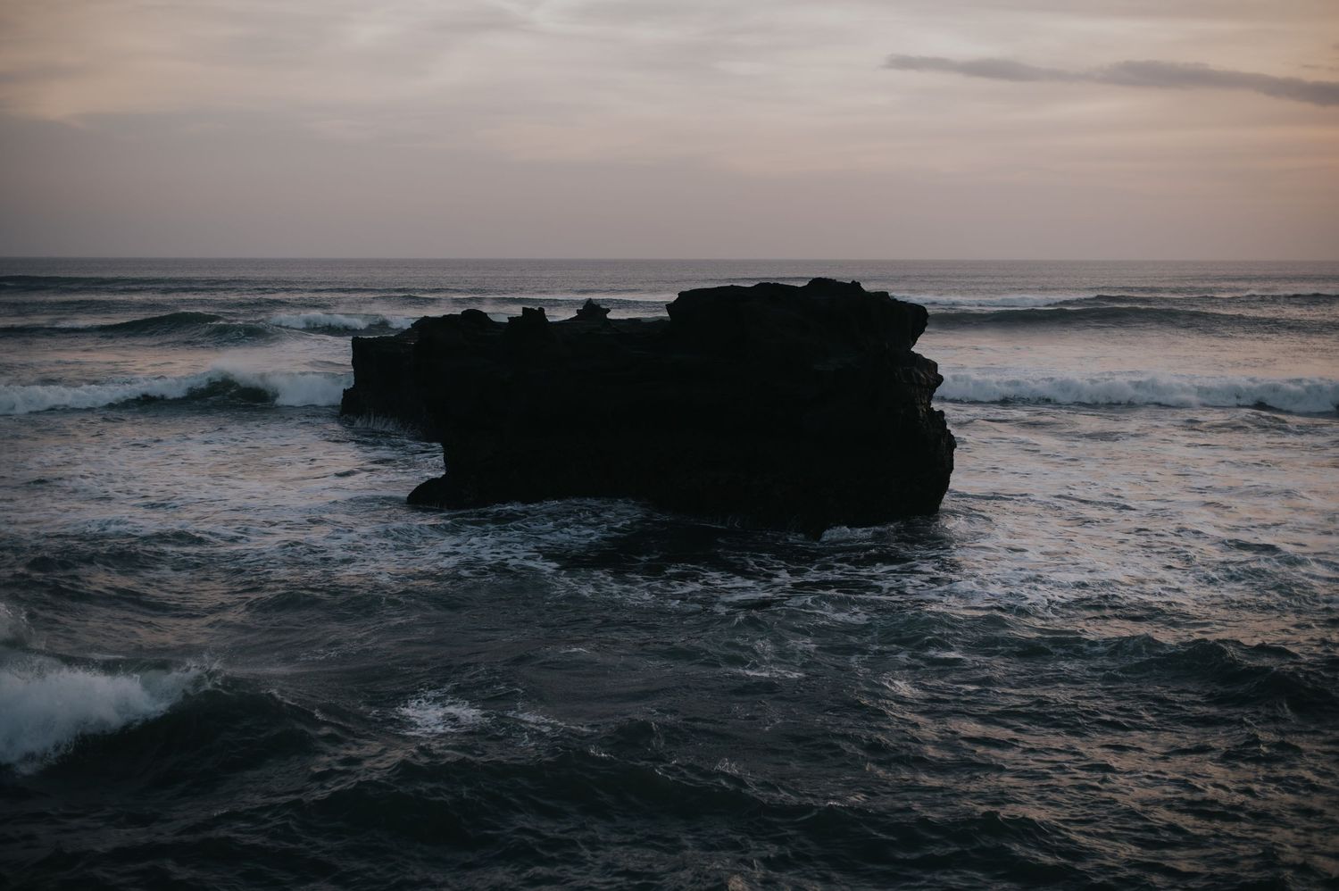 Dark rocky outcrop silhouetted against moody ocean waves and cloudy sunset sky.