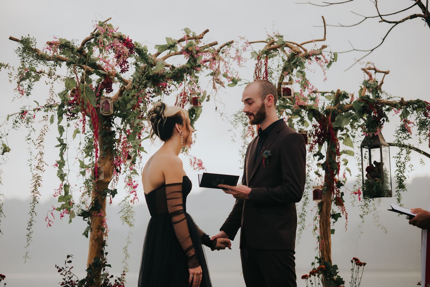 A couple stands beneath an arch decorated with red flowers and greenery during a ceremony.