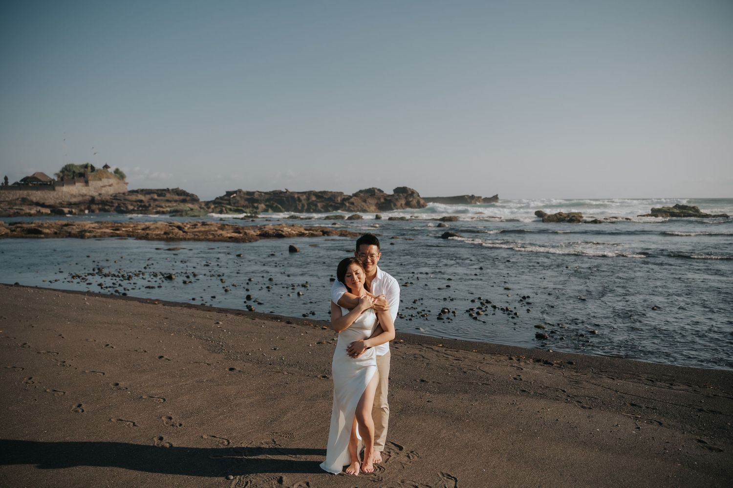 Couple embracing on a sandy beach with rocky coastline and ocean waves in the background during golden hour.