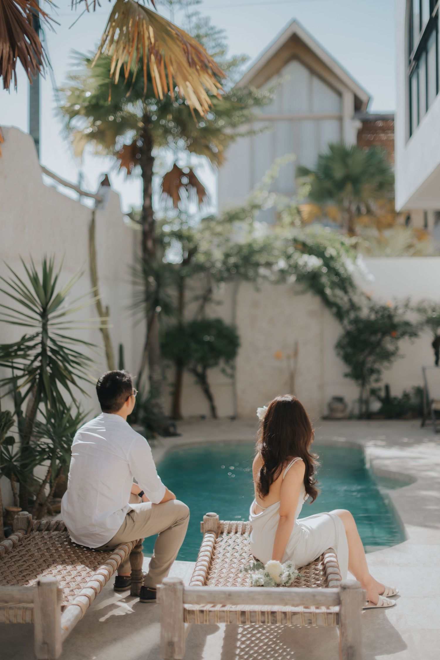 Couple in white attire sitting by a tropical pool surrounded by lush greenery and modern villa architecture.
