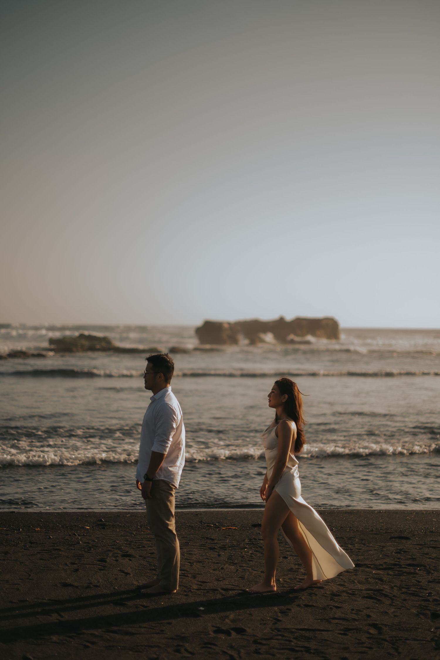 Couple walking on a sandy beach at sunset with ocean waves and a rocky outcrop in the background.