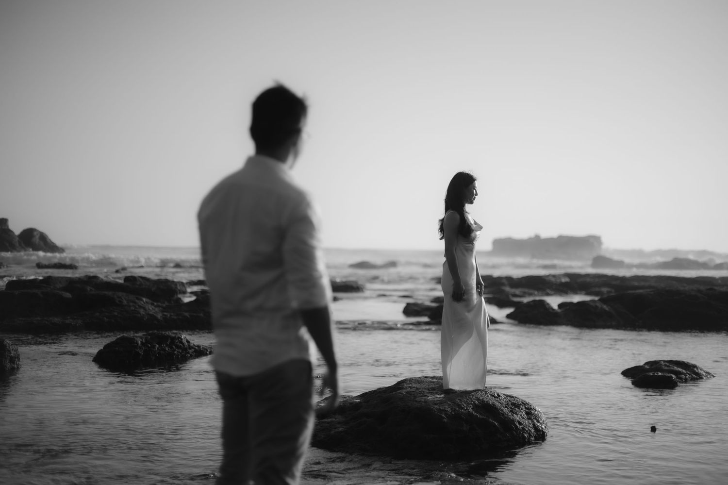 Black and white photo of a couple on rocky beach, woman in white dress stands on rock as man watches from foreground.