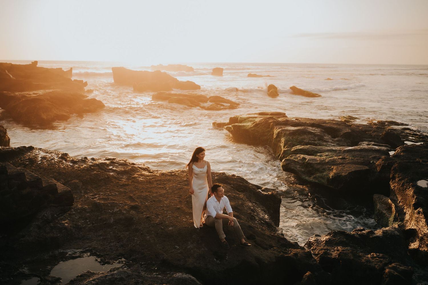 Couple posing on rocky coastal cliffs at golden sunset with ocean waves in background.