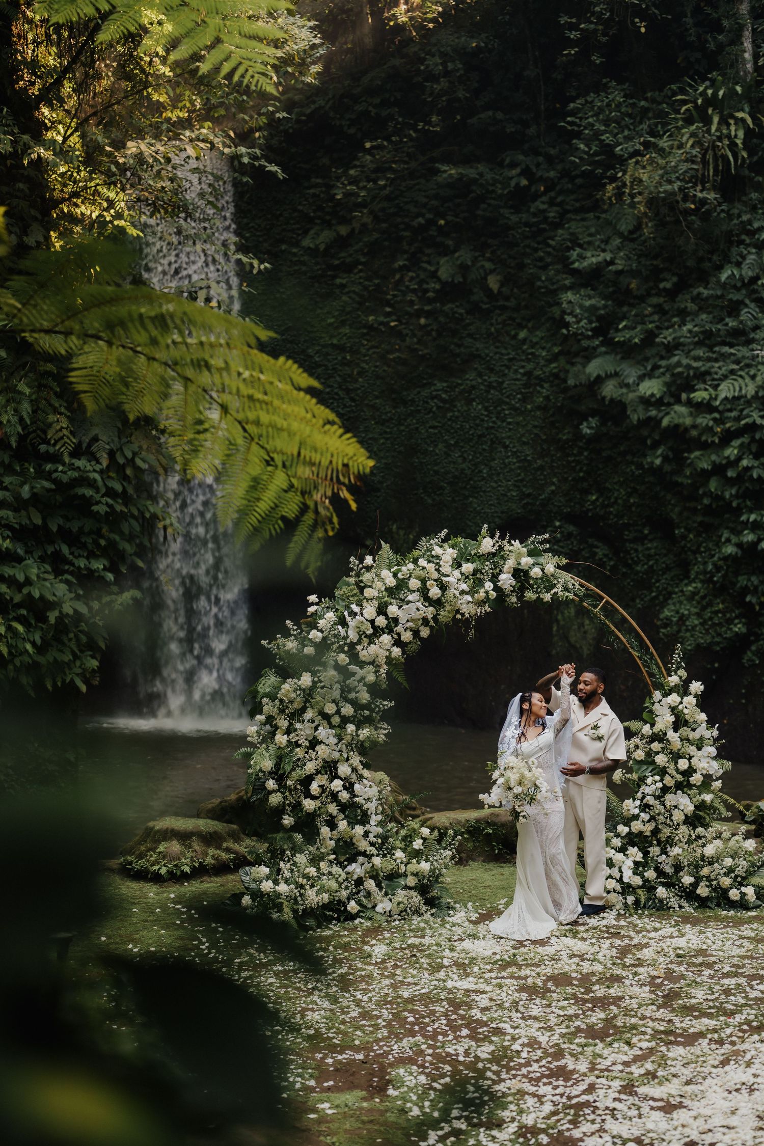 Bride and groom posing under white flower arch surrounded by lush greenery and waterfall.