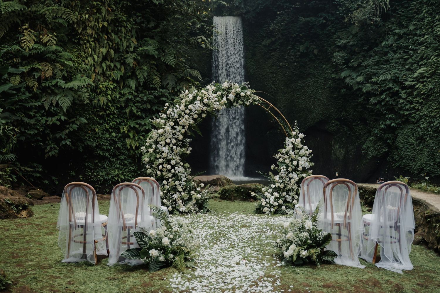 Romantic outdoor wedding ceremony setup with floral arch and waterfall backdrop in lush green forest.