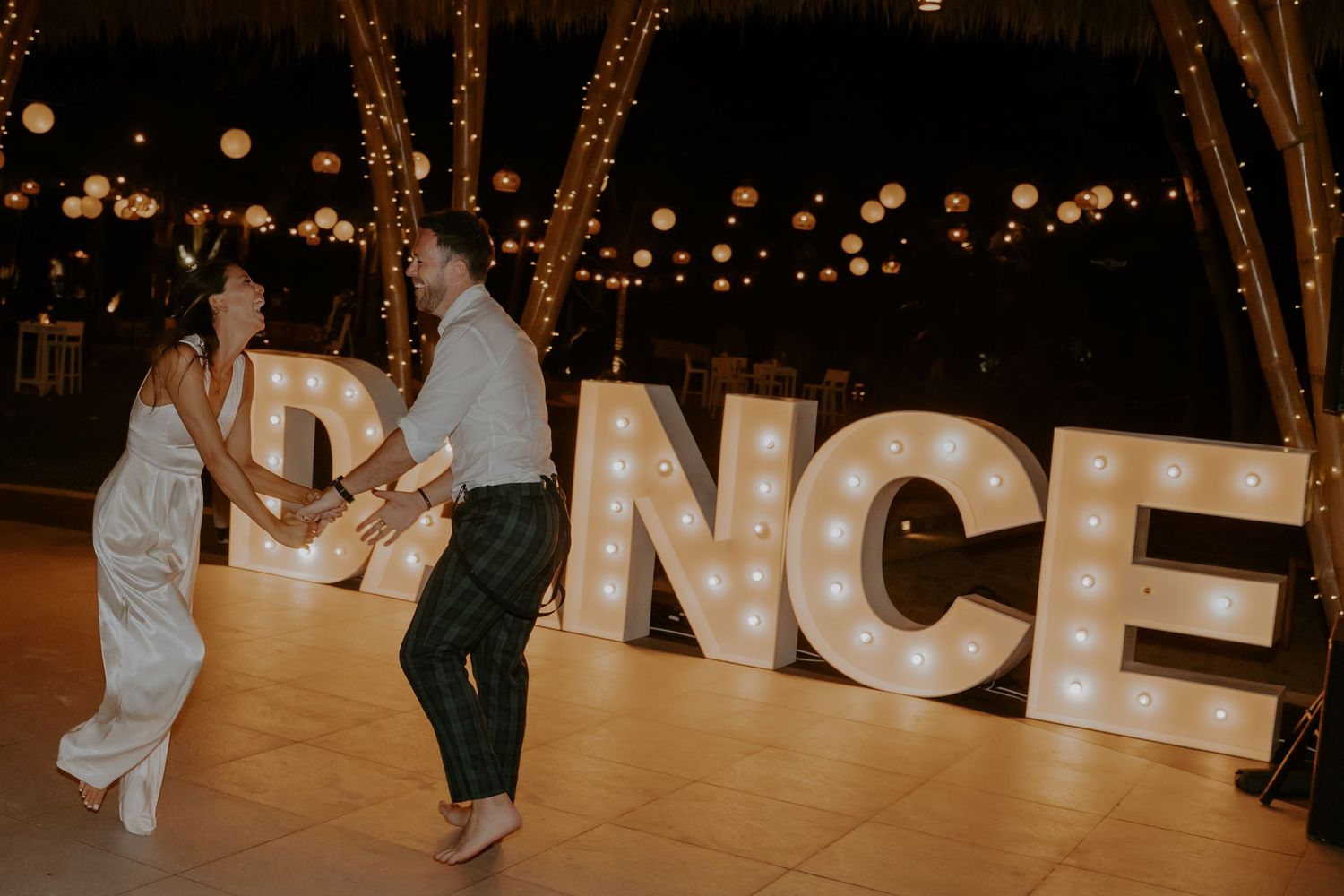 A romantic couple dances together under twinkling lights at an outdoor evening wedding reception.