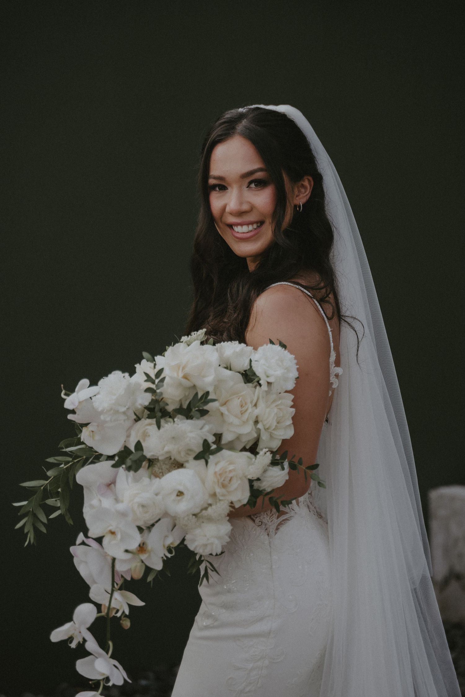 A bride in a white wedding dress holds a cascading bouquet of white orchids and roses against a dark background.
