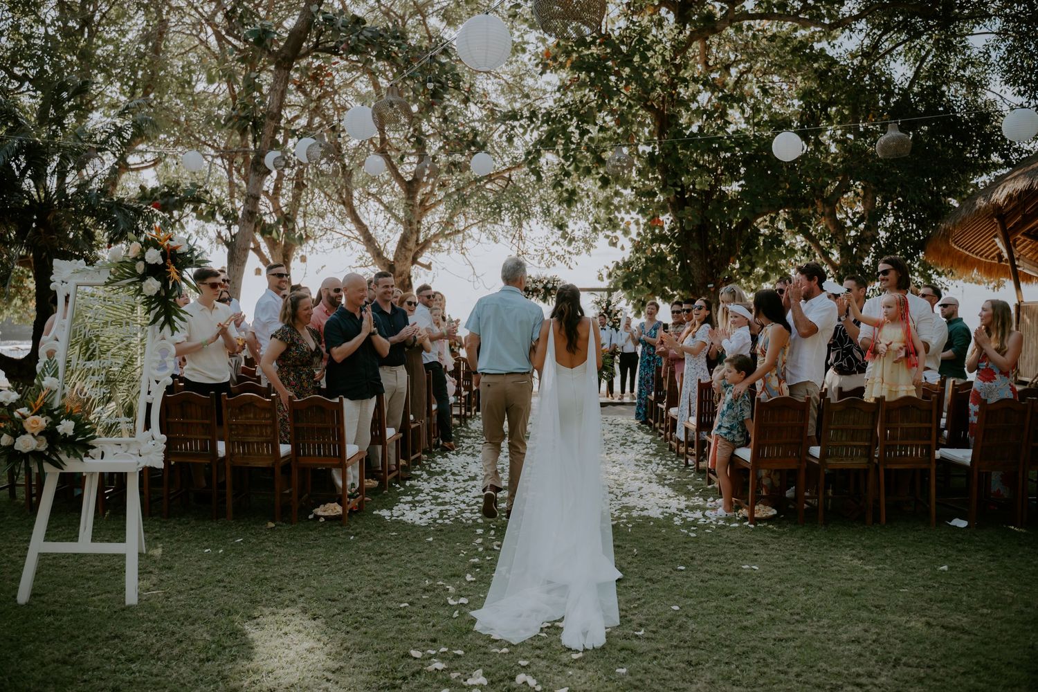 Wedding guests gather outdoors under trees at a rustic ceremony venue during sunset.