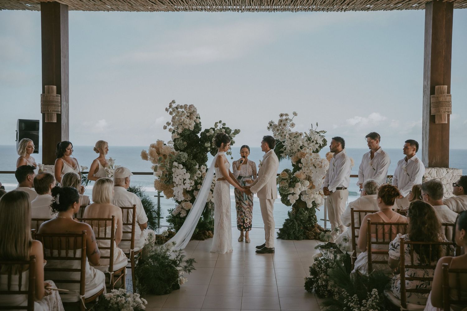 Wedding guests seated at long tables during an outdoor reception with ocean views and tropical landscaping.