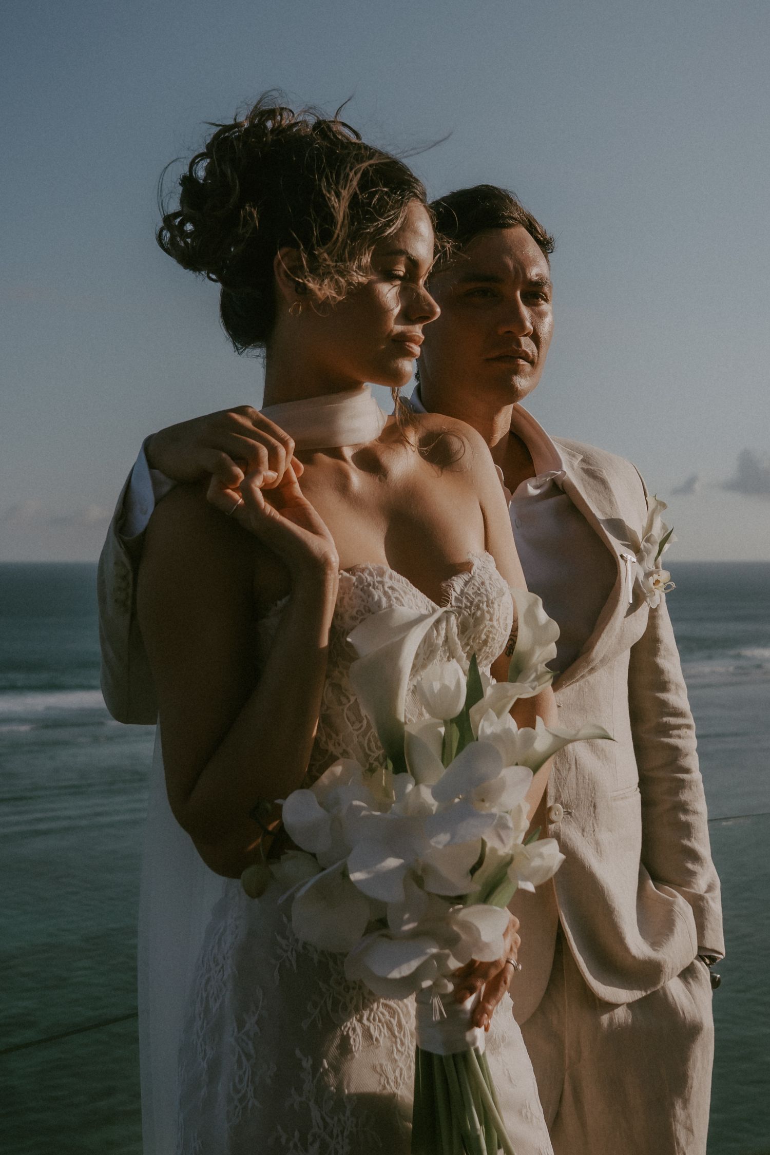 Romantic coastal wedding portrait with couple embracing by ocean at sunset in elegant white and beige attire.