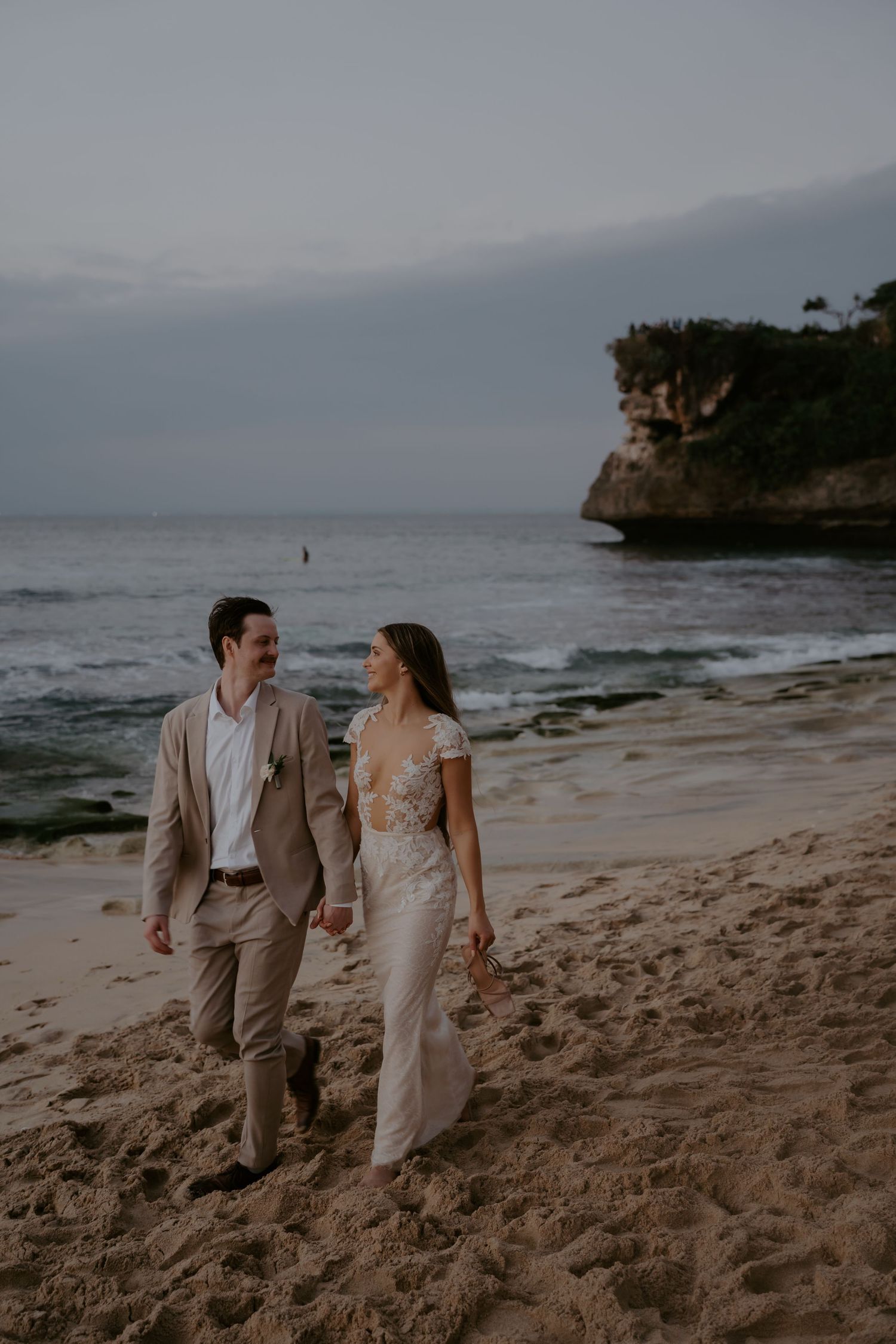 A couple in light-colored attire walking hand in hand along a scenic beach with cliffs in the background at sunset.