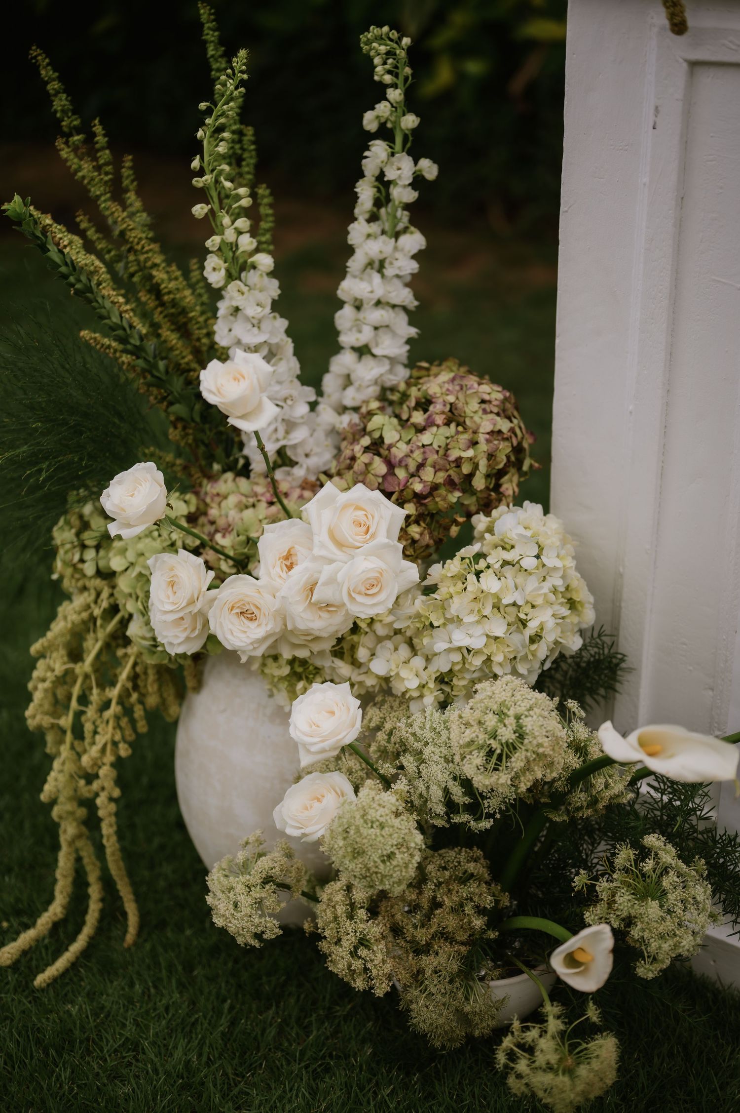 Elegant white floral arrangement with roses, hydrangeas, and delphinium in a stone vase at an outdoor wedding ceremony.