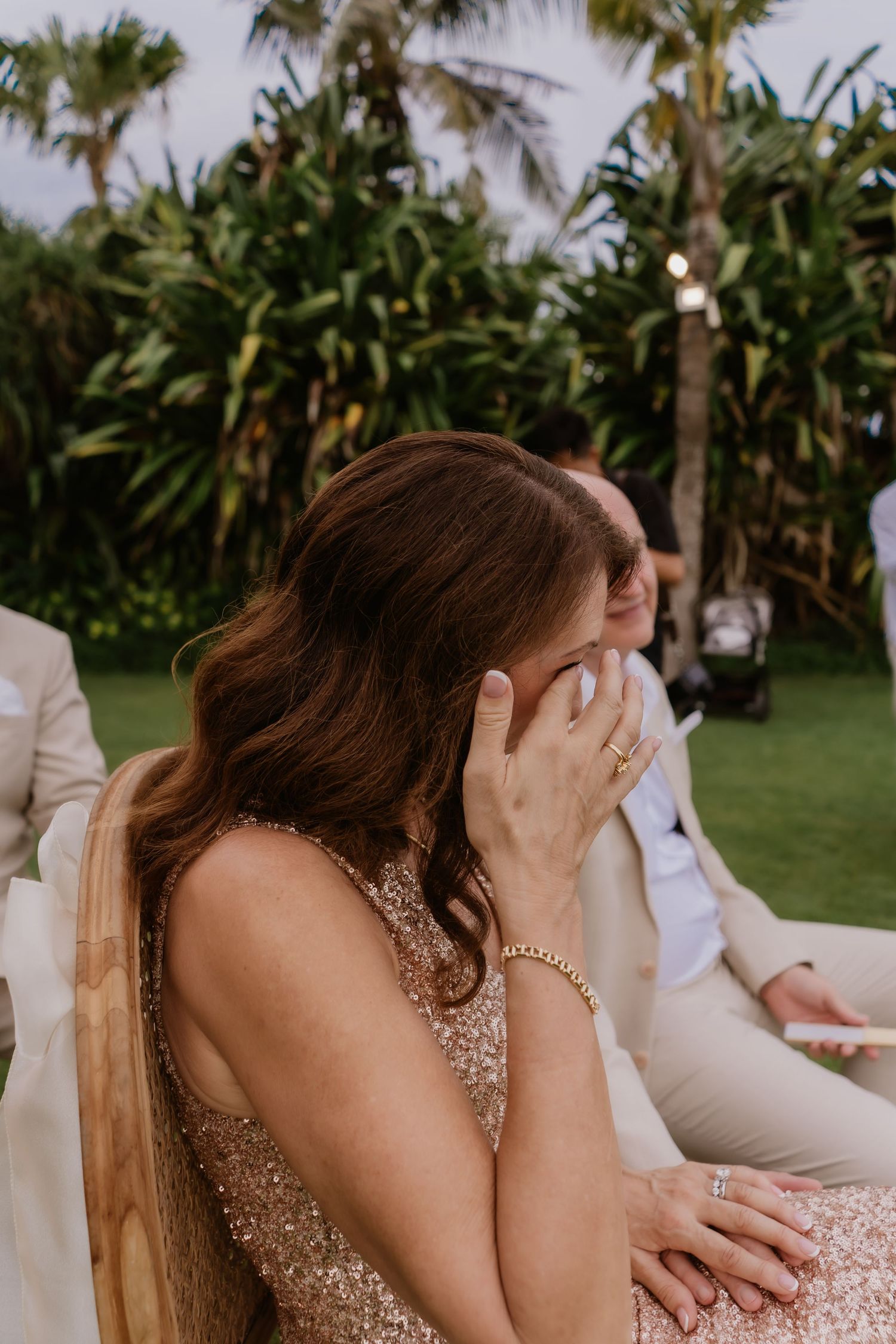 Emotional wedding guest in gold sequin dress wiping tears during outdoor ceremony with tropical greenery.
