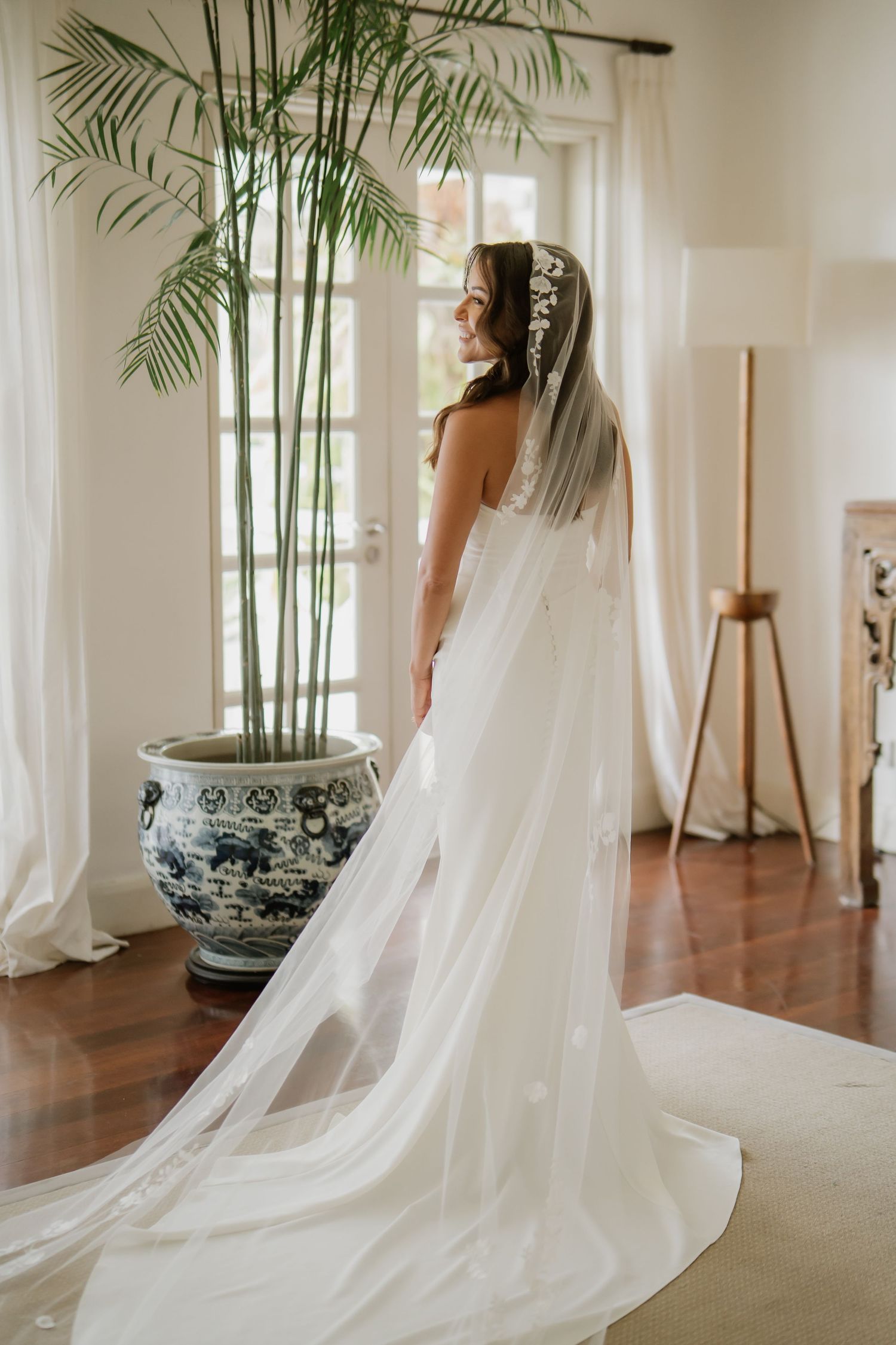 A bride in a flowing white gown and long veil stands by a window in elegant decor.