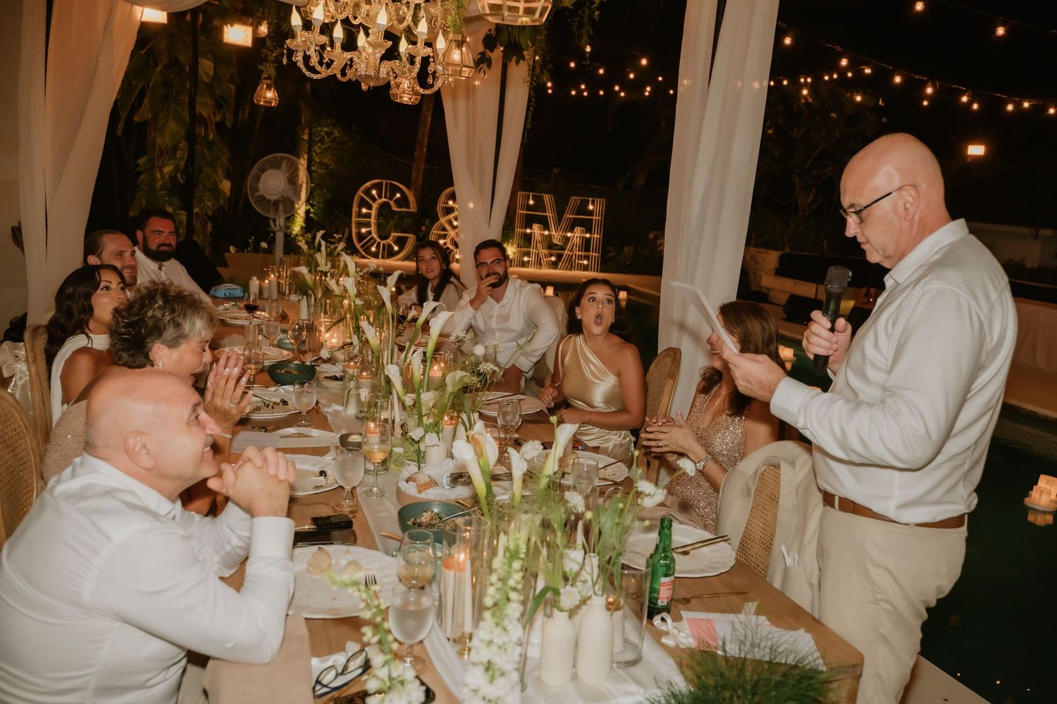Wedding reception dinner with guests applauding a speech under chandelier and string lights.