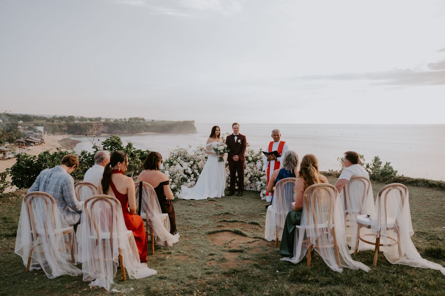Intimate outdoor wedding ceremony overlooking ocean cliffs with guests seated in rattan chairs around couple.