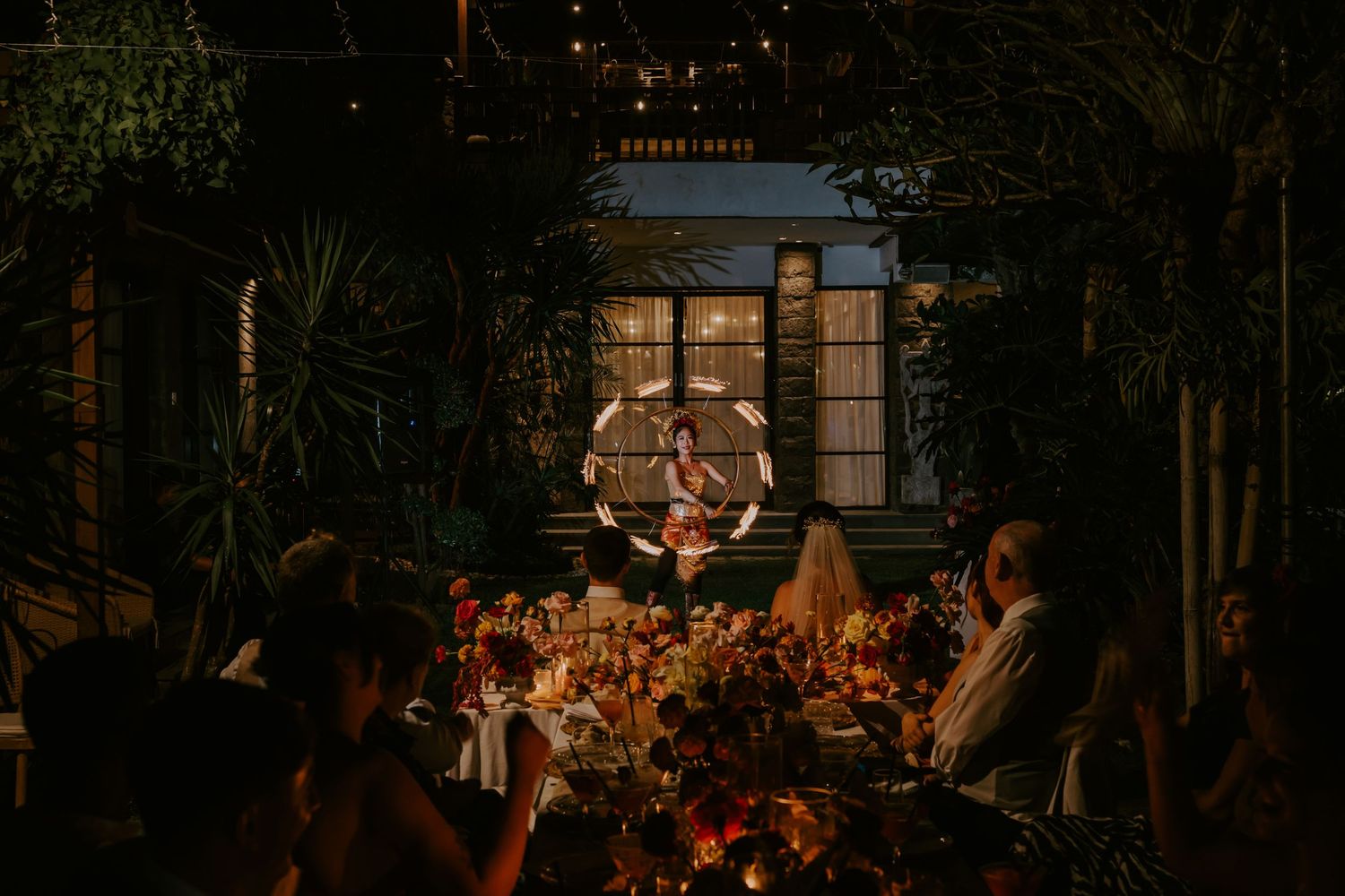 Wedding reception guests dine as hoop performer entertains under warm glowing lights amid tropical decor.