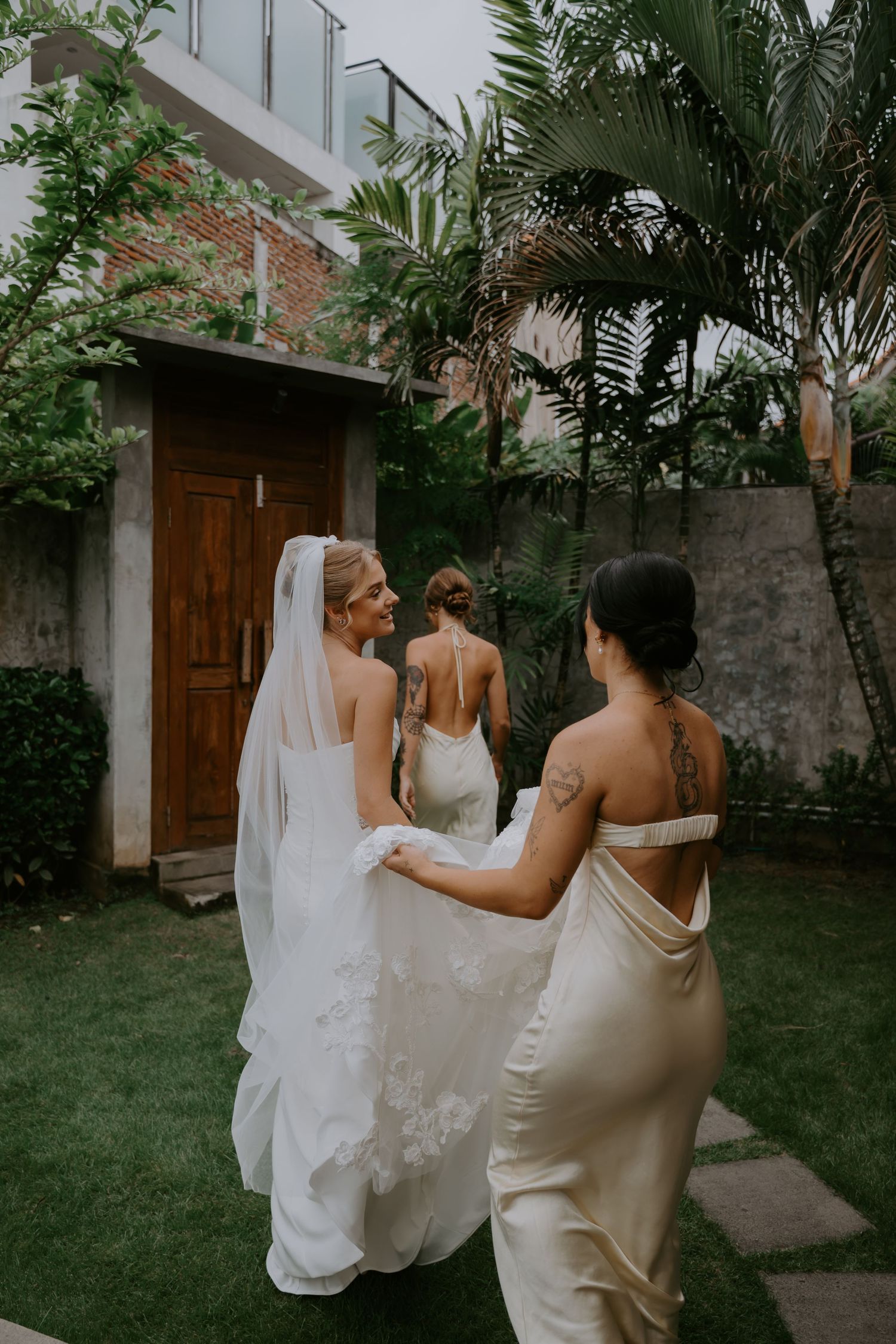 Bridesmaid holds bride's lace wedding gown train in lush tropical garden setting.