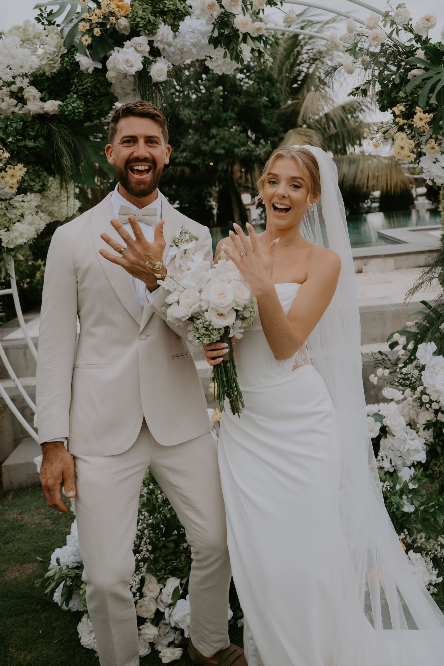 Newlyweds joyfully show wedding rings in front of white floral arch after outdoor ceremony.