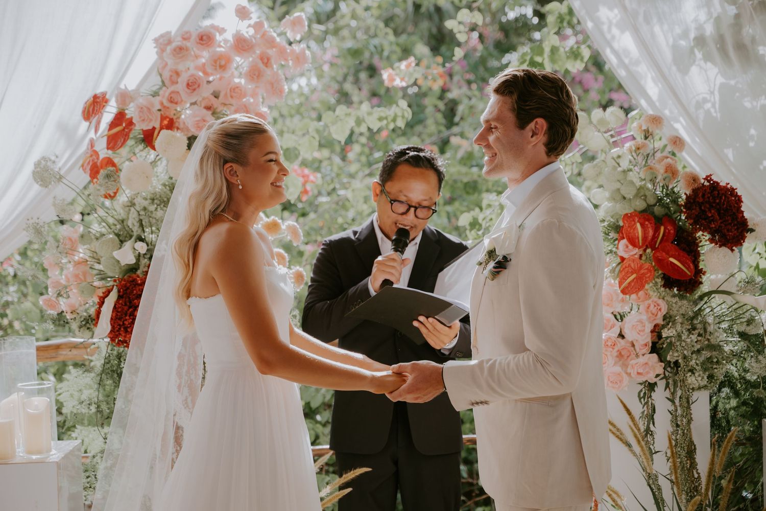 Bride and groom exchange vows before officiant beneath a stunning floral arch of pink and red blooms.