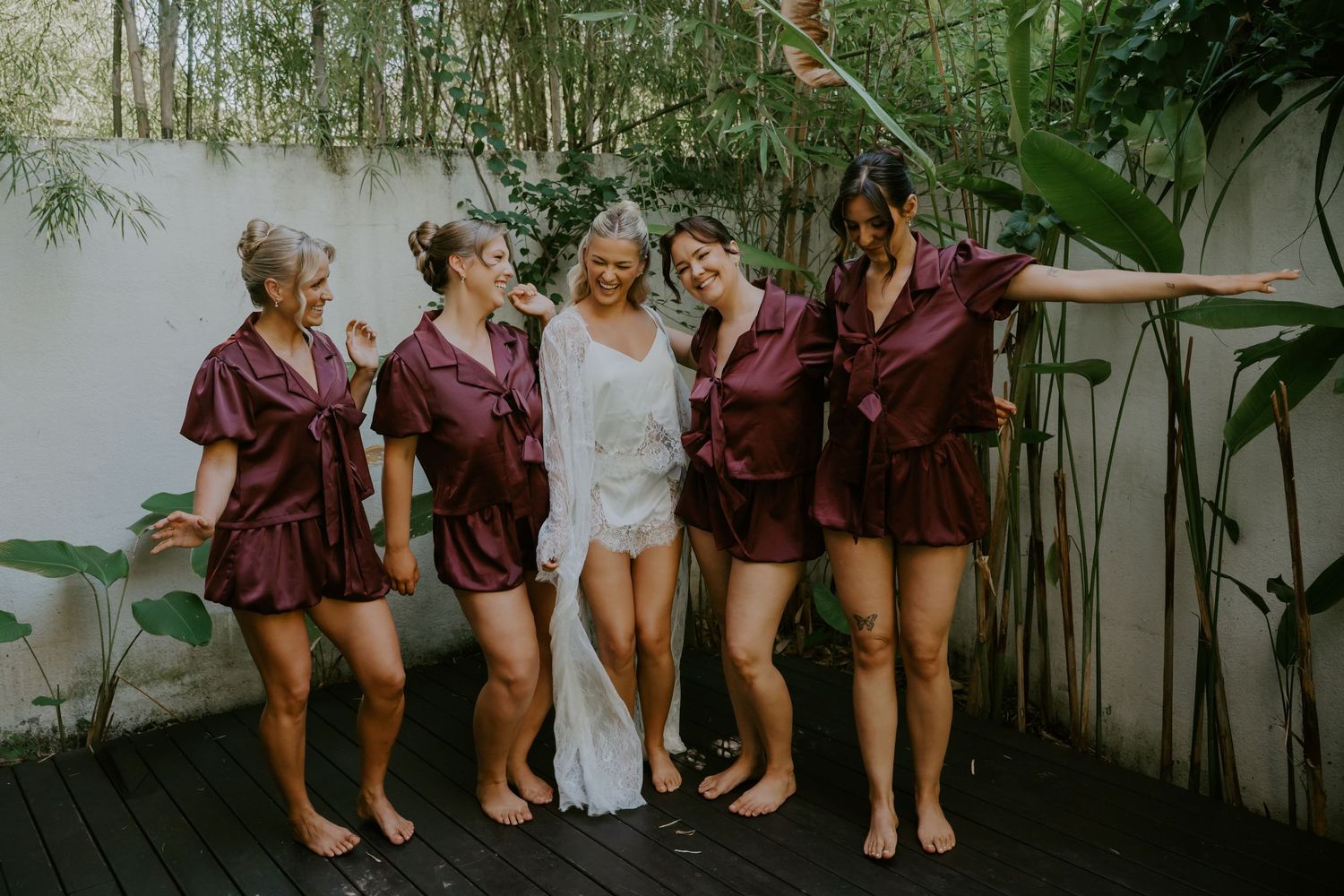 Bride in white lace robe poses with four bridesmaids in matching burgundy satin pajama sets on a wooden deck.