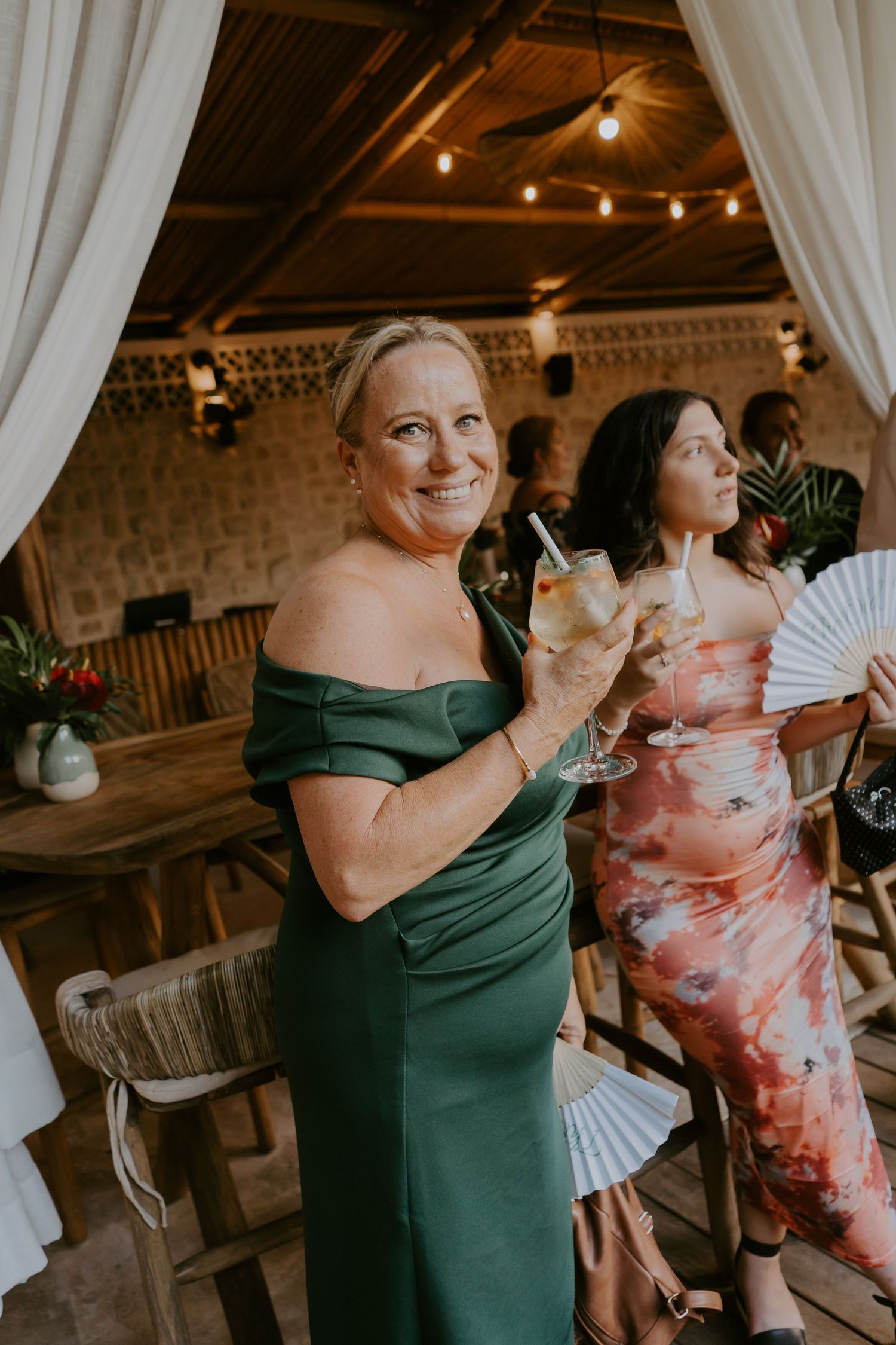 Elegant woman in off-shoulder green gown holds cocktail at tropical wedding venue.