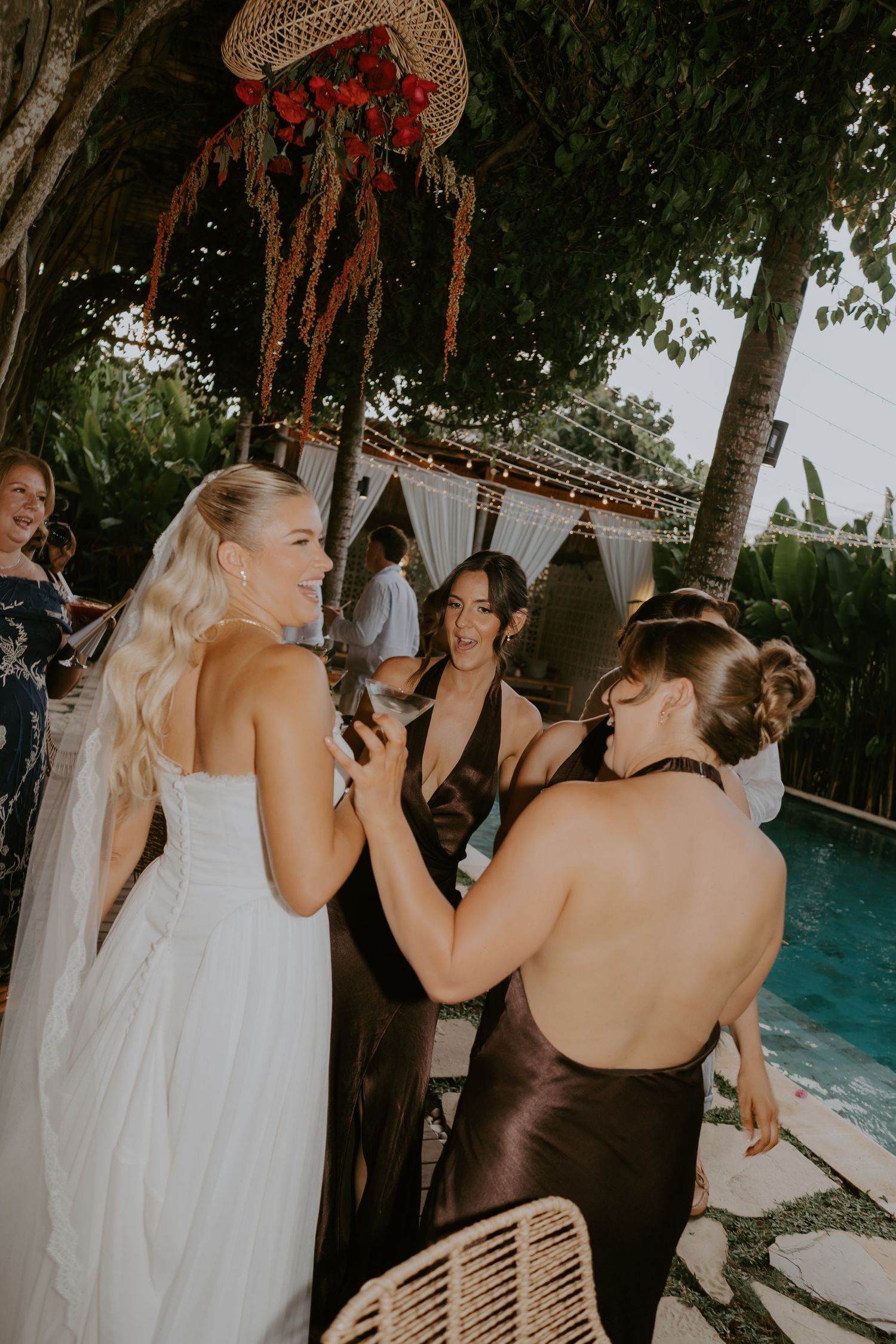 Bride in white strapless gown laughing with bridesmaids in black dresses at outdoor poolside reception.