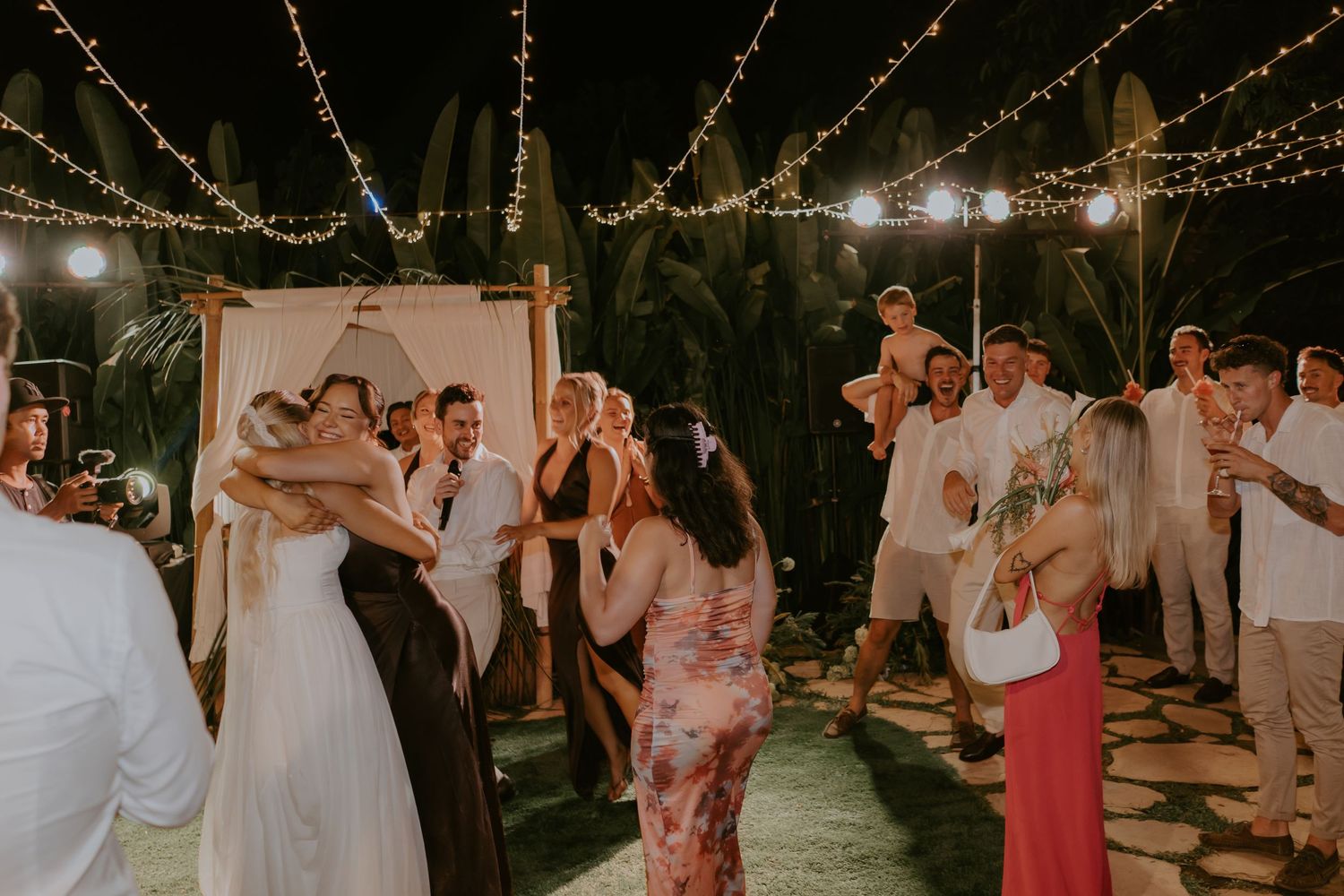 Bride and guests dance joyfully under string lights at tropical outdoor wedding reception at night.