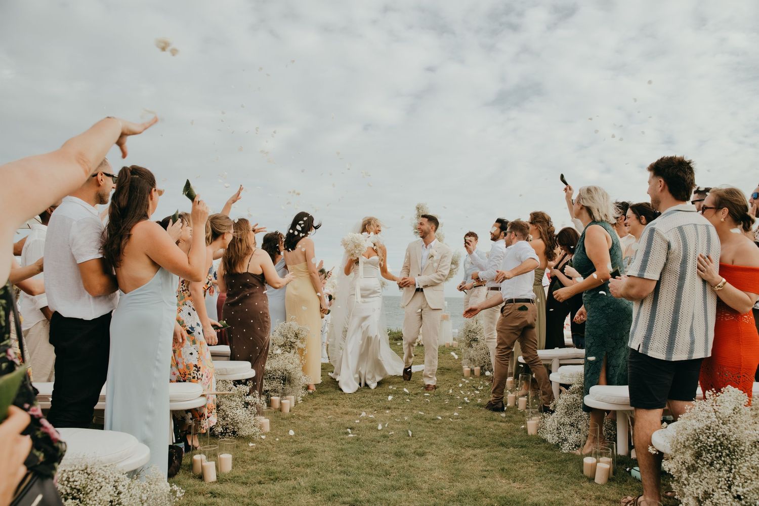 Newlyweds walk down aisle as guests toss petals in celebration at outdoor tropical wedding ceremony.