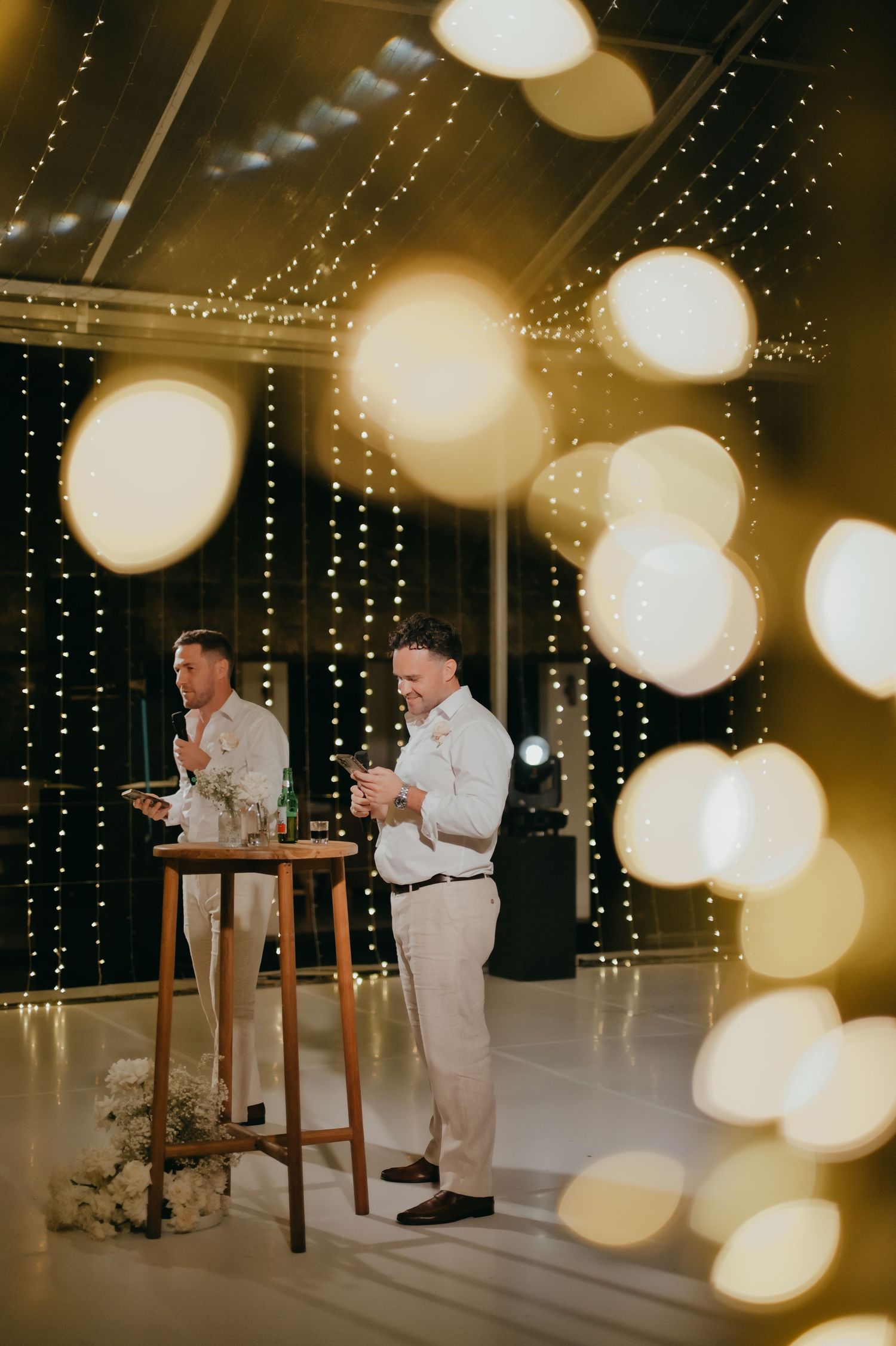 Two groomsmen giving speeches at a wedding reception surrounded by warm fairy lights.