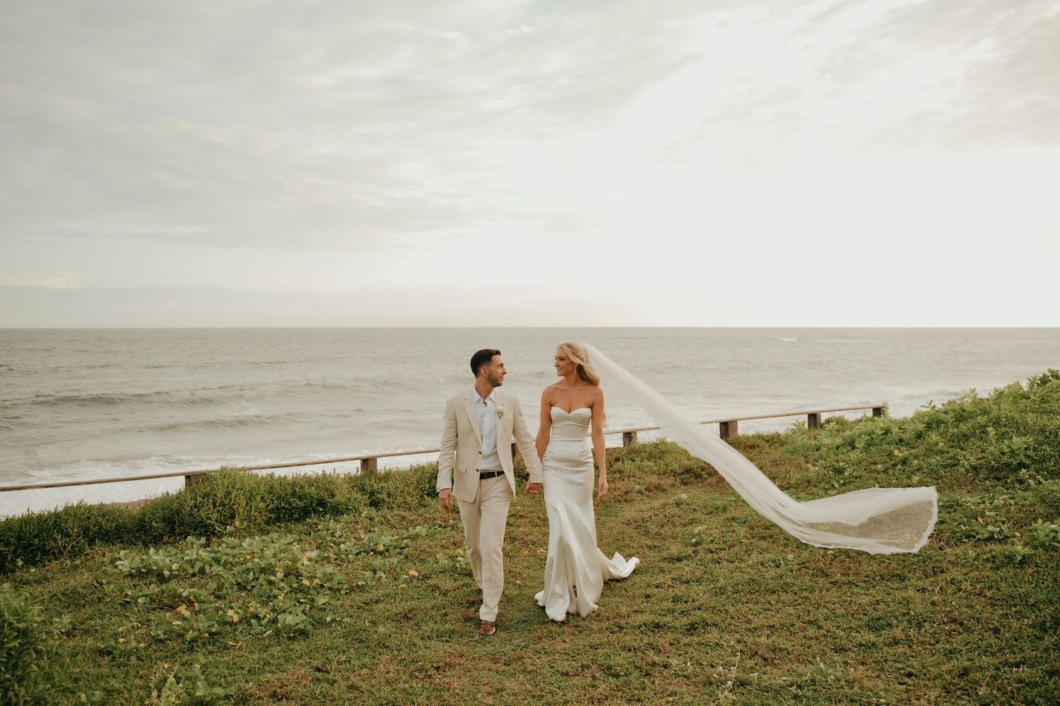 Bride and groom walk hand-in-hand on a grassy cliff overlooking the ocean, veil blowing in the wind.