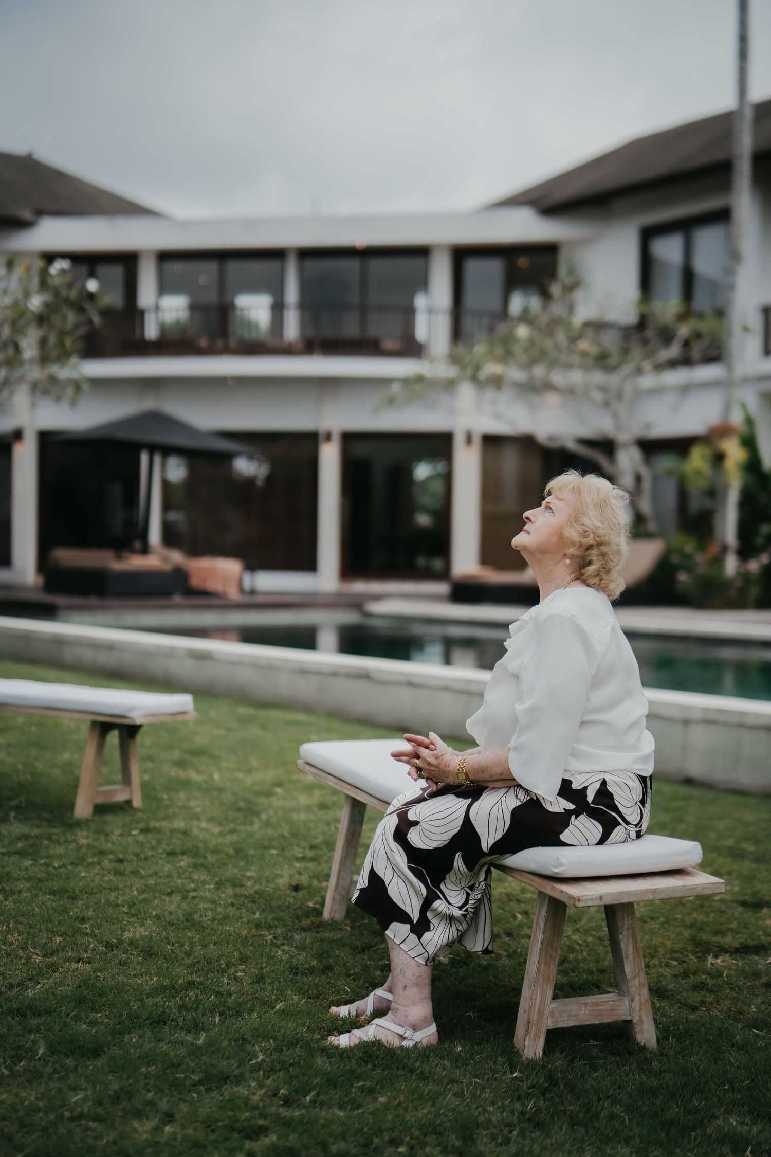 Elegant older woman sitting on a wooden bench outdoors, laughing joyfully beside a luxury villa pool.