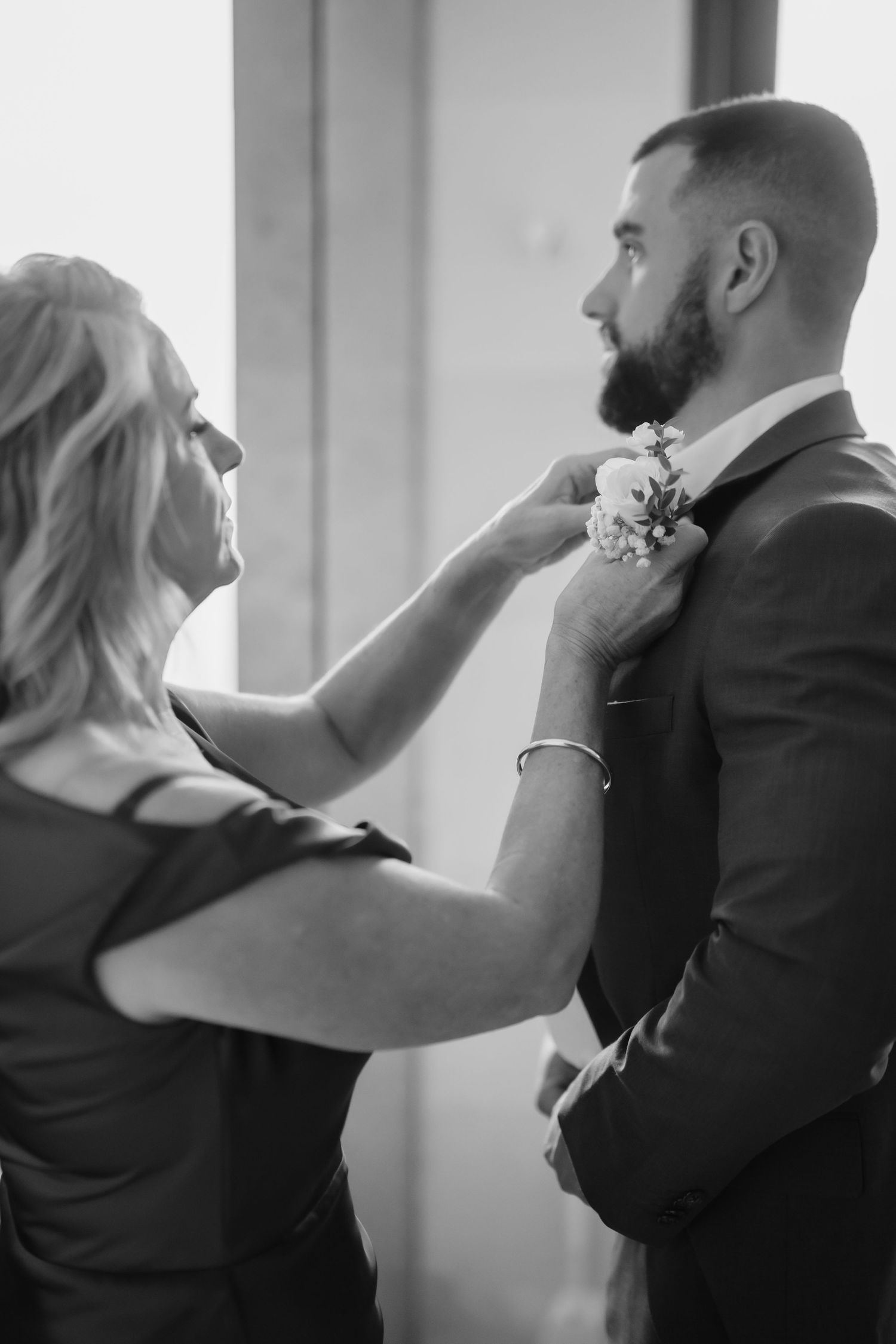 Black and white photo of a woman pinning a floral boutonniere on a bearded groom before the wedding.
