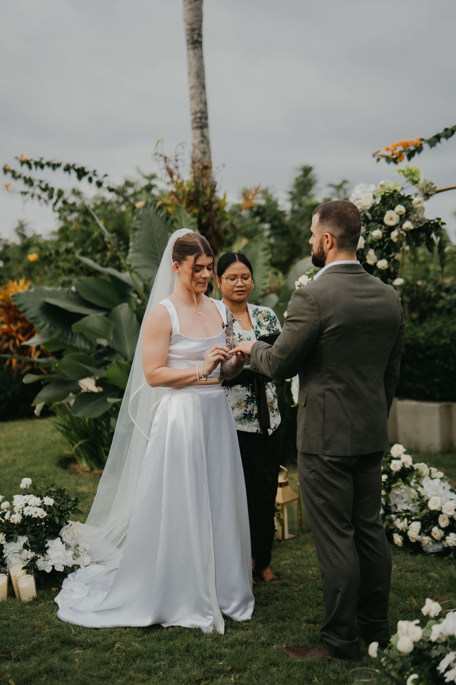 Bride and groom exchange rings during outdoor garden ceremony with white floral arch and officiant in Bali.