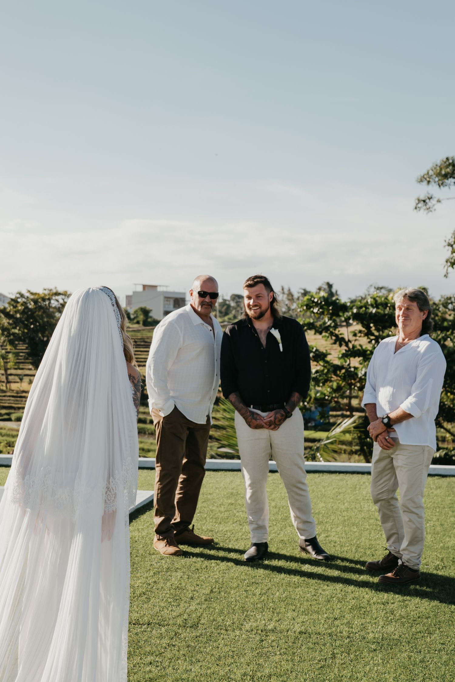 Bride in white veil walks toward groom and guests during outdoor wedding ceremony on green lawn.