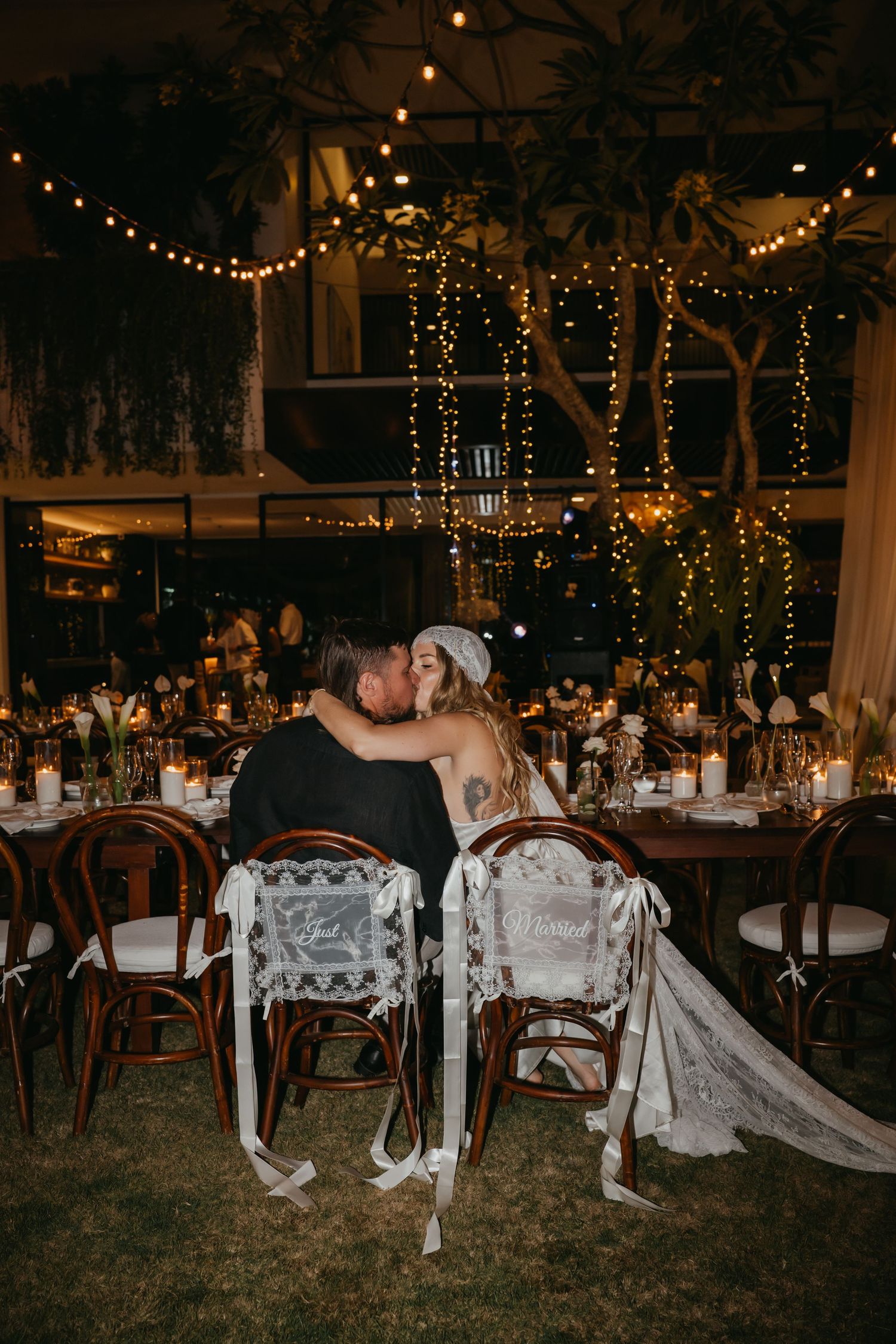 Bride and groom share a kiss at their candlelit outdoor wedding reception table adorned with fairy lights.