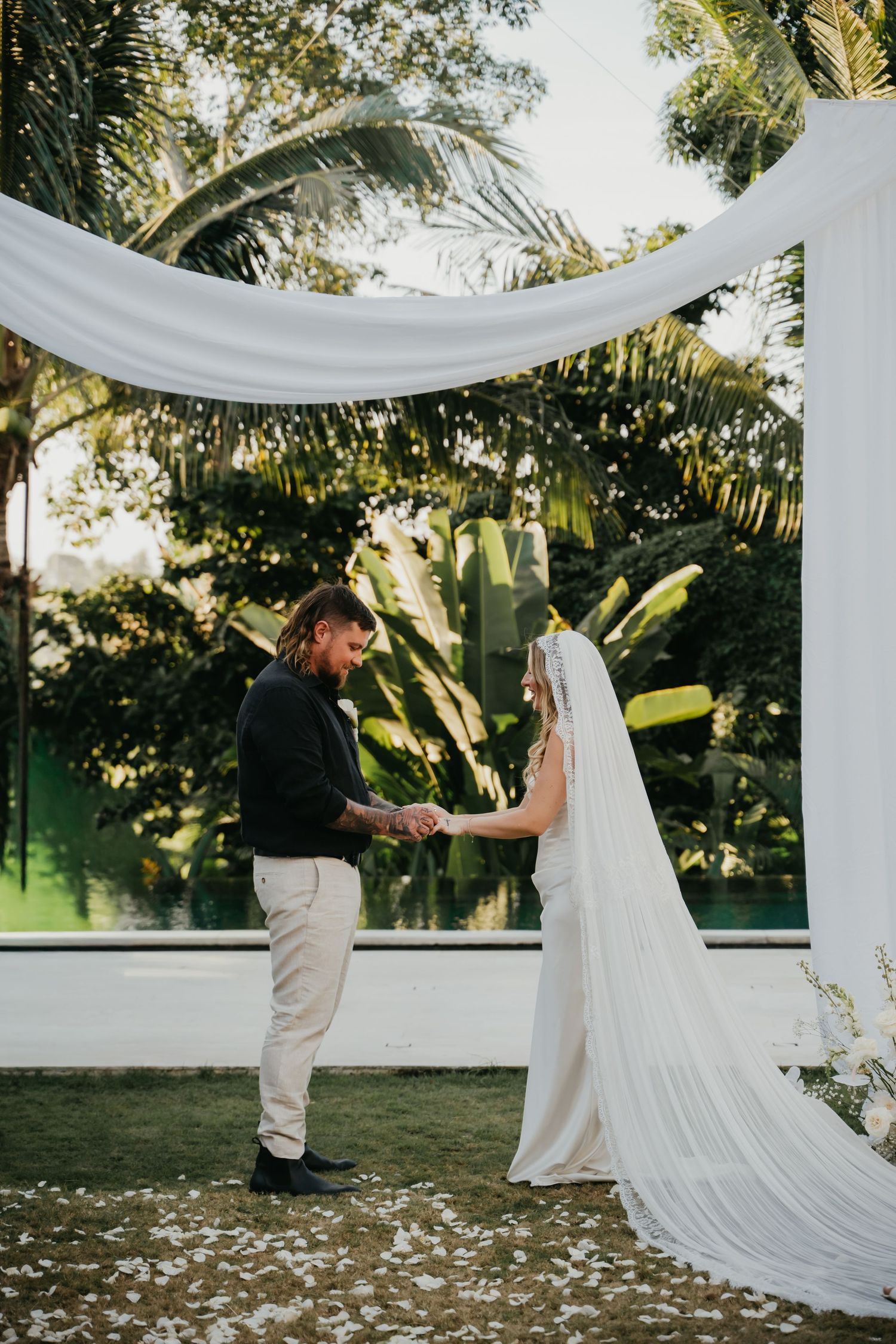 Bride and groom exchange vows under a white draped arch surrounded by tropical greenery.
