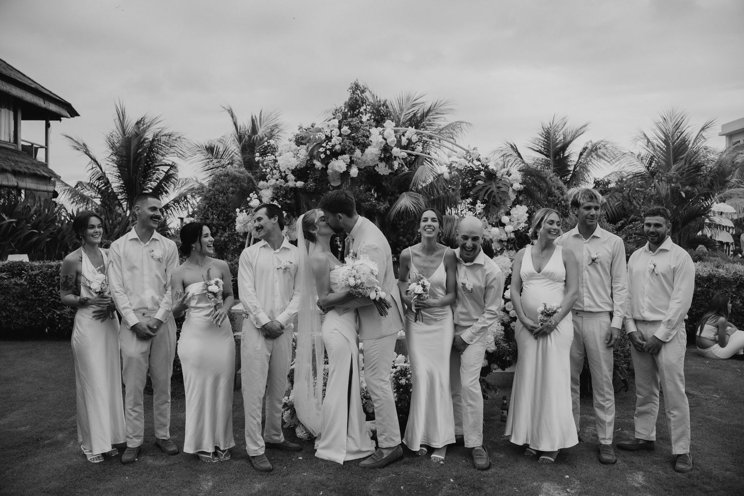Black and white wedding party photo with bride and groom kissing, flanked by bridesmaids and groomsmen, tropical palm trees backdrop.