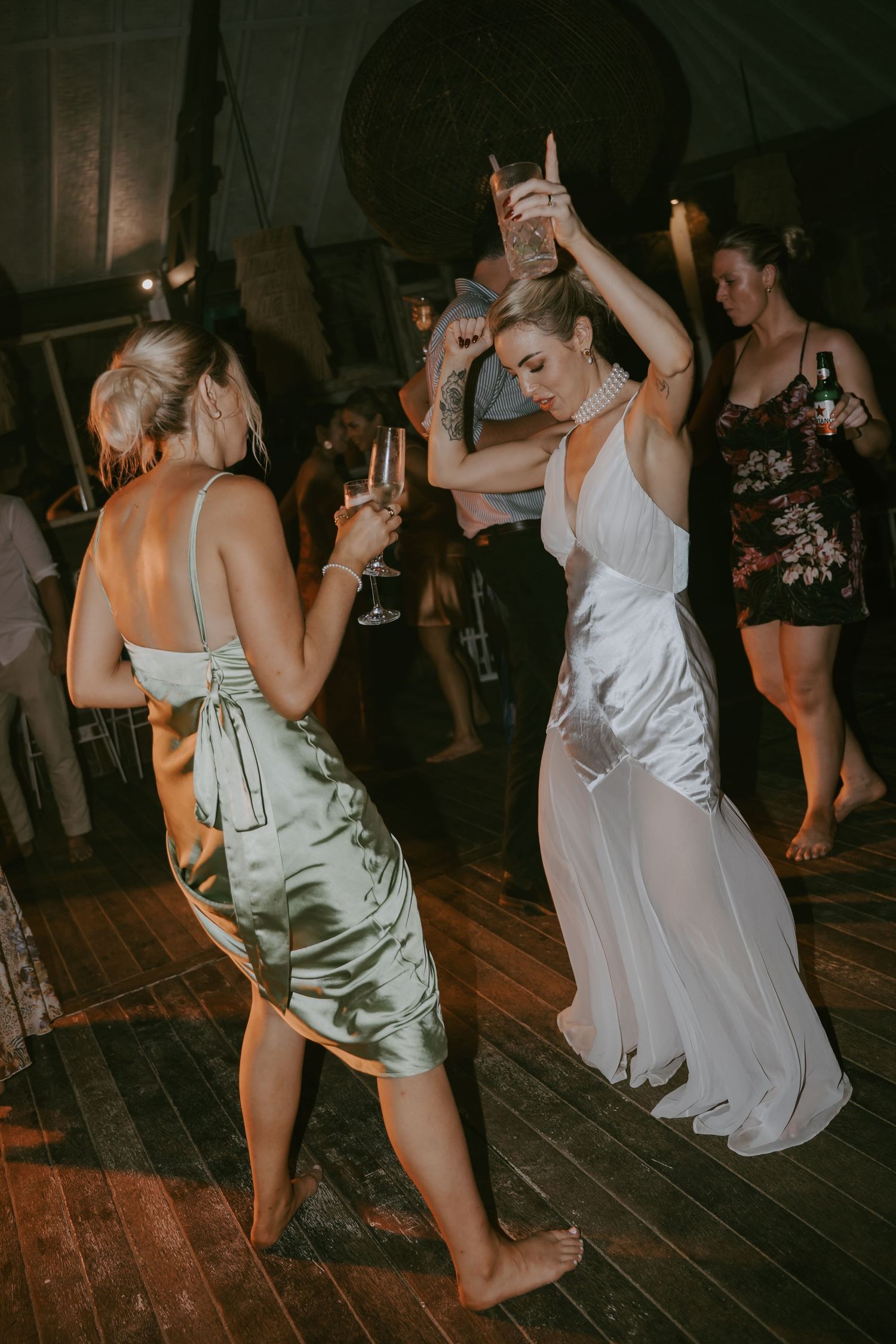 Wedding guests dancing barefoot on a wooden dance floor in elegant dresses at a dimly lit reception.