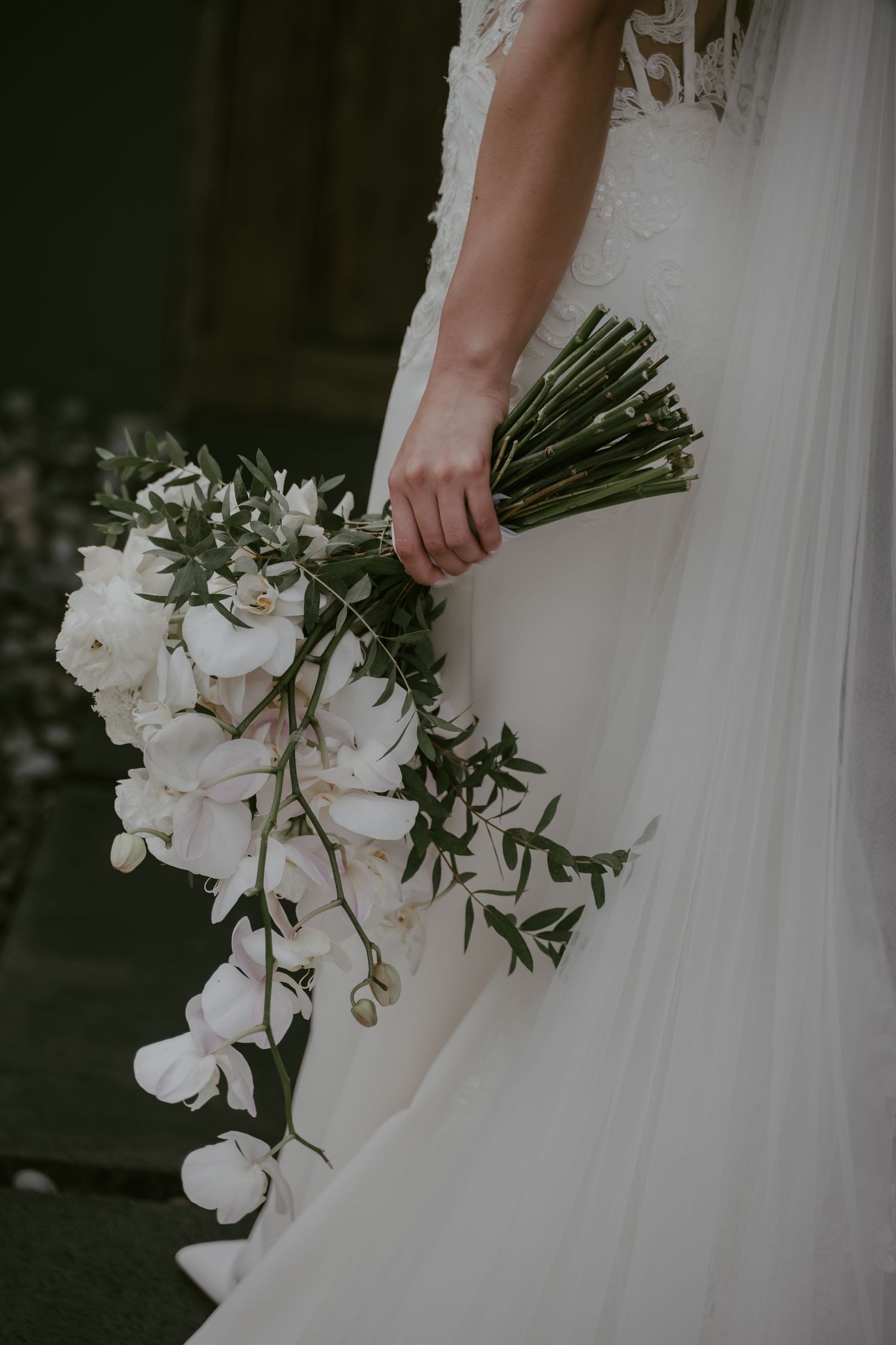 A cascading white orchid bouquet held against a flowing white wedding dress in a moody, dark setting.