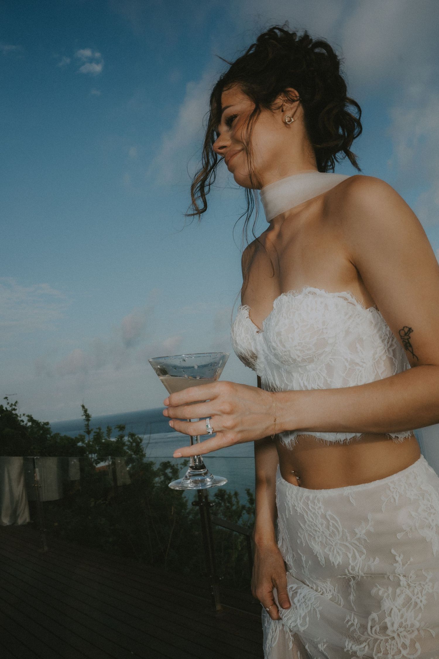 A figure in a white lace wedding dress stands against a scenic tropical backdrop with blue skies and ocean views.